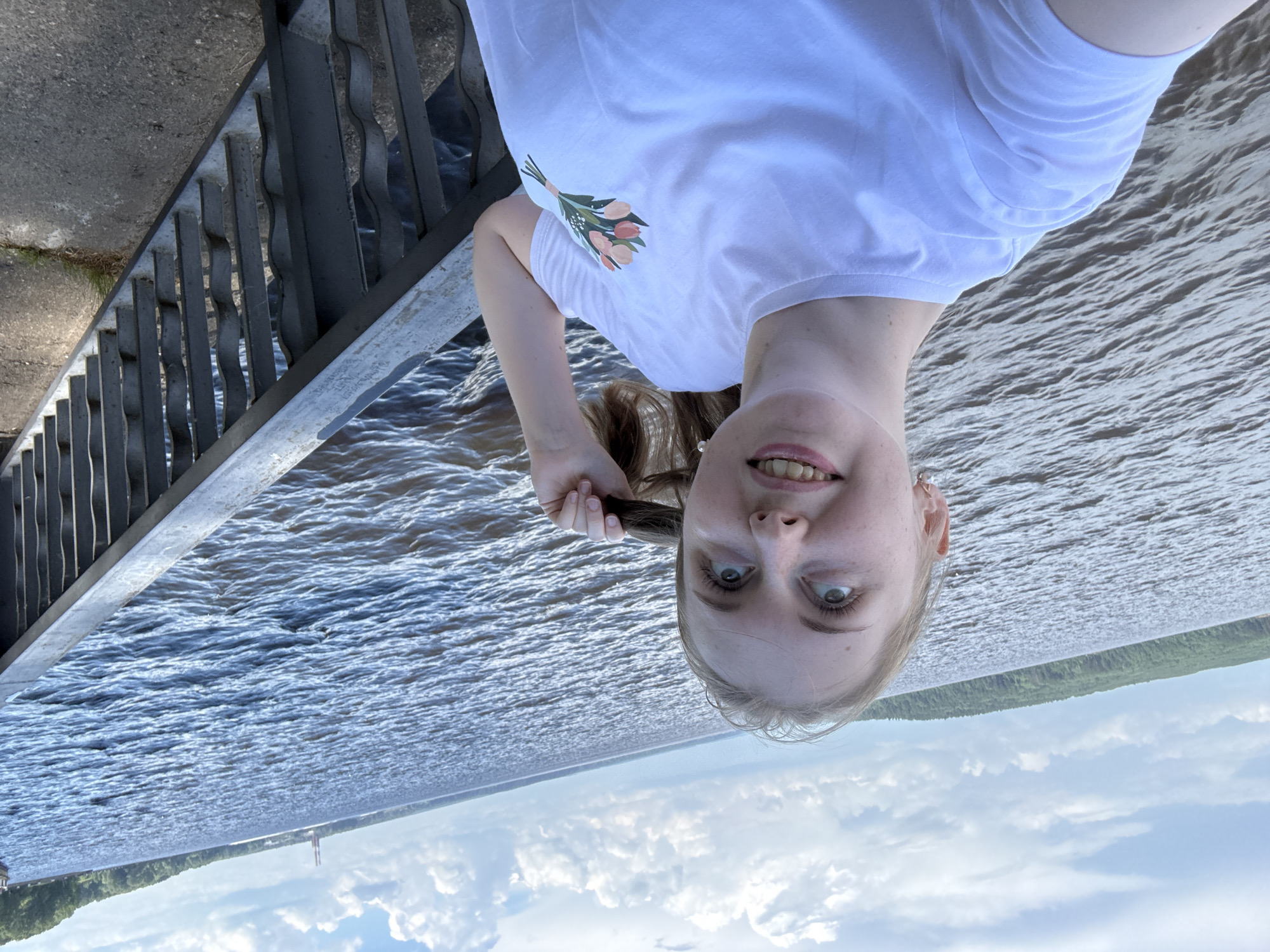 A young woman with blonde hair stands on a paved area next to a railing, looking up and to her right. She is wearing a white t-shirt with a floral print on the left side of her chest. Her hair is partially pulled back, and she is holding a strand of it. The railing is made of dark metal with decorative vertical bars. Beyond the railing is a wide body of water with gentle waves, and in the distance, a tree-covered shoreline is visible. The sky is partly cloudy with white clouds against a light blue background. The overall scene appears to be outdoors on a bright day, possibly along a waterfront promenade or park.