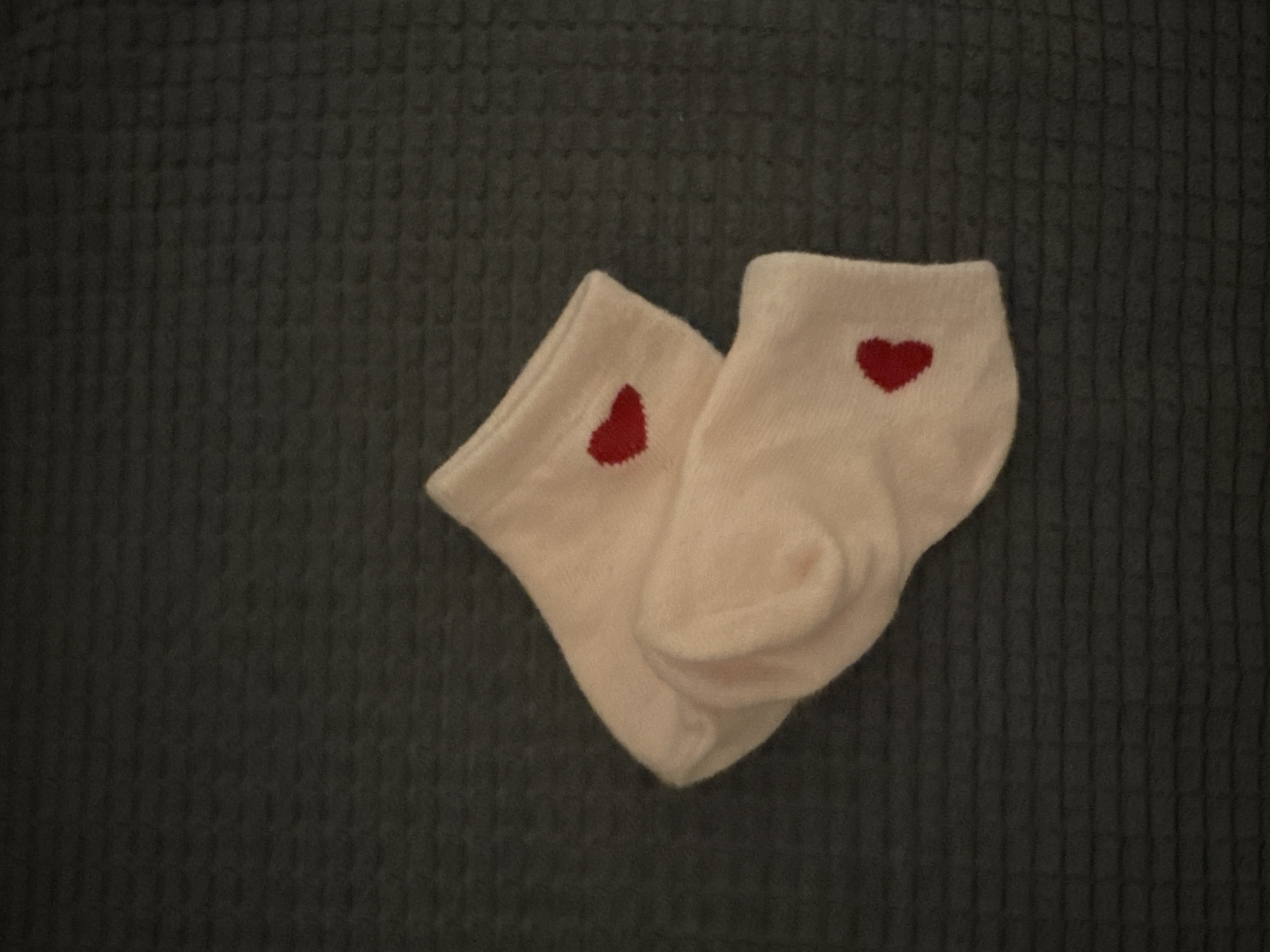 Two pink socks with red heart appliques are lying on a dark, textured surface. One sock is layered on top of the other. The socks appear to be made of a soft material, possibly wool or a blend. The background is dark gray with a subtle grid-like pattern. The lighting is dim, casting soft shadows. No people are visible in the image. The location is unknown.
