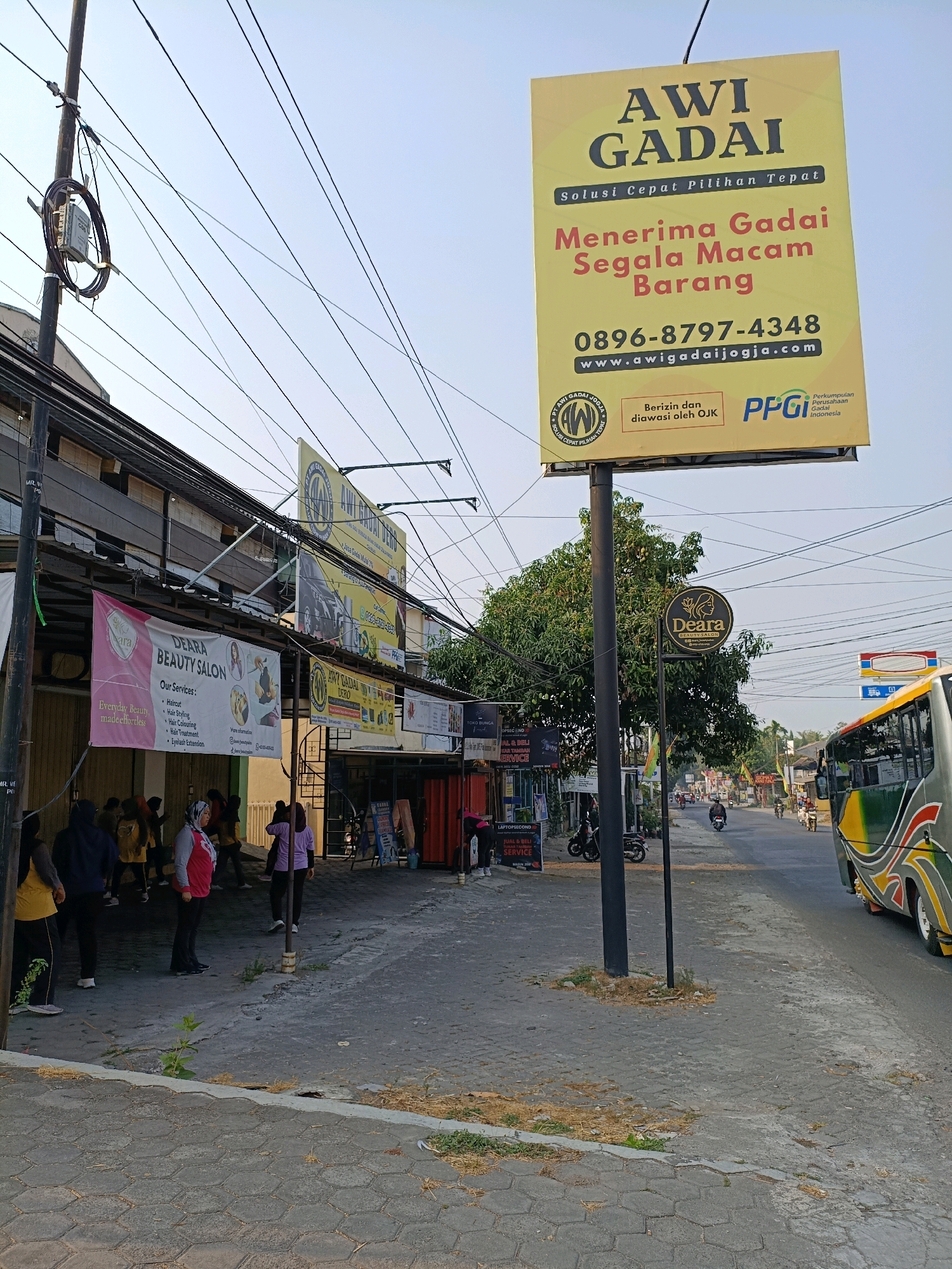 The image depicts a street scene in Ngemplak, Indonesia, likely during daytime with a clear sky. A prominent yellow billboard with the text "AWI GADAI" and "Menerima Gadai Segala Macam Barang" is a central focus, advertising a pawn shop. Below this, contact information and a website address are visible. To the left of the billboard, a banner for "DEARA BEAUTY SALON" is displayed. Several people are gathered on the sidewalk in front of the establishments, some appearing to be engaged in an activity. A black pole supports the billboard. The street is paved, and a bus is visible on the right side of the road, along with other vehicles and motorbikes further down the street. Overhead, numerous electrical wires crisscross the scene, typical of urban infrastructure. The overall environment suggests a commercial area with multiple businesses.