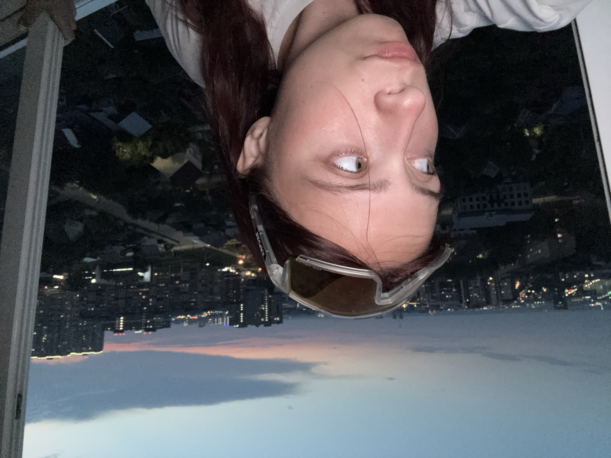 The image shows a young woman with auburn hair wearing large, clear-lensed sunglasses on her forehead. She is positioned in the foreground, facing left and looking out of frame, with a contemplative expression. Her head and shoulders are visible, and she appears to be leaning against a white window frame.
The background depicts a sprawling city skyline at dusk. The sky is a gradient of deep blue to soft pink and orange, indicating the transition from daylight to night. Numerous buildings of various heights are visible, some with illuminated windows, suggesting an urban environment. The scene appears to be a residential area with houses and apartment buildings.
The overall setting is an apartment window in Izhevsk, Russia, offering a panoramic view of the city as the day concludes.