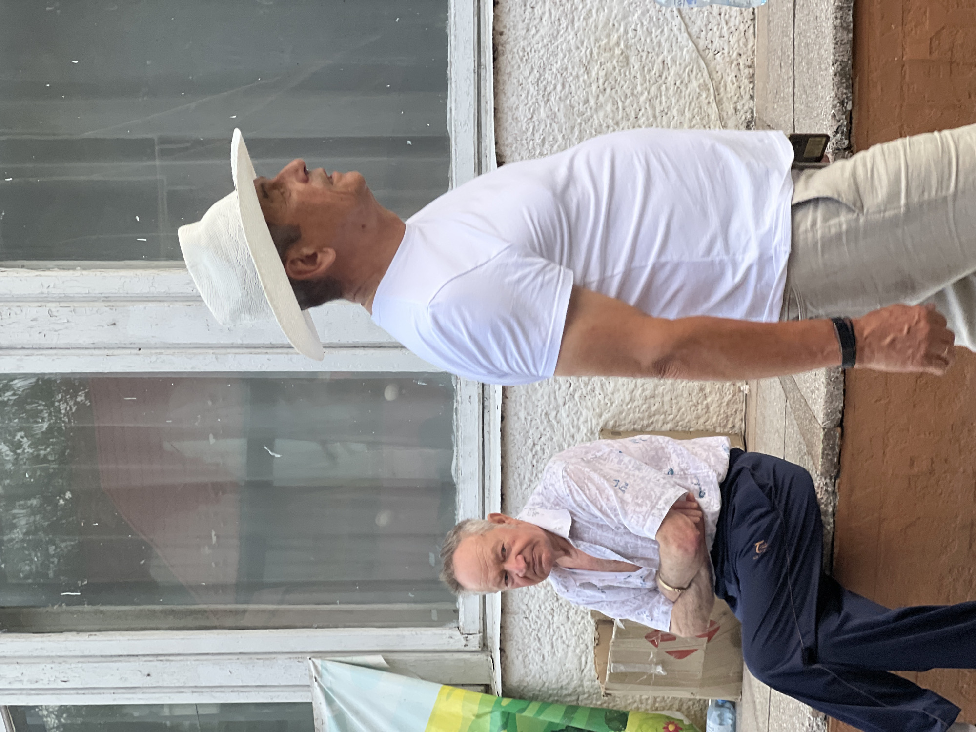 The image depicts an outdoor scene on a daytime, possibly late afternoon, based on the lighting. Two middle-aged to older Caucasian men are the primary subjects. In the foreground, on the right, a man wearing a white t-shirt and a wide-brimmed white hat is seen in profile, facing right and appearing to walk or stand. He has a noticeably larger belly. To his left, further in the background and seated on a low ledge, is another man with a fair complexion, grey hair, and a somewhat displeased expression. He is wearing a light-colored patterned short-sleeved shirt with unbuttoned top buttons and dark blue sweatpants. His arms are crossed. Behind the seated man, there are cardboard boxes. The background shows a building with a window, the glass of which reflects the surroundings. The environment appears to be urban or suburban, with a textured wall of the building. No specific landmarks or indicators of the city of Сонячне, Ukraine are present in the visual information.
