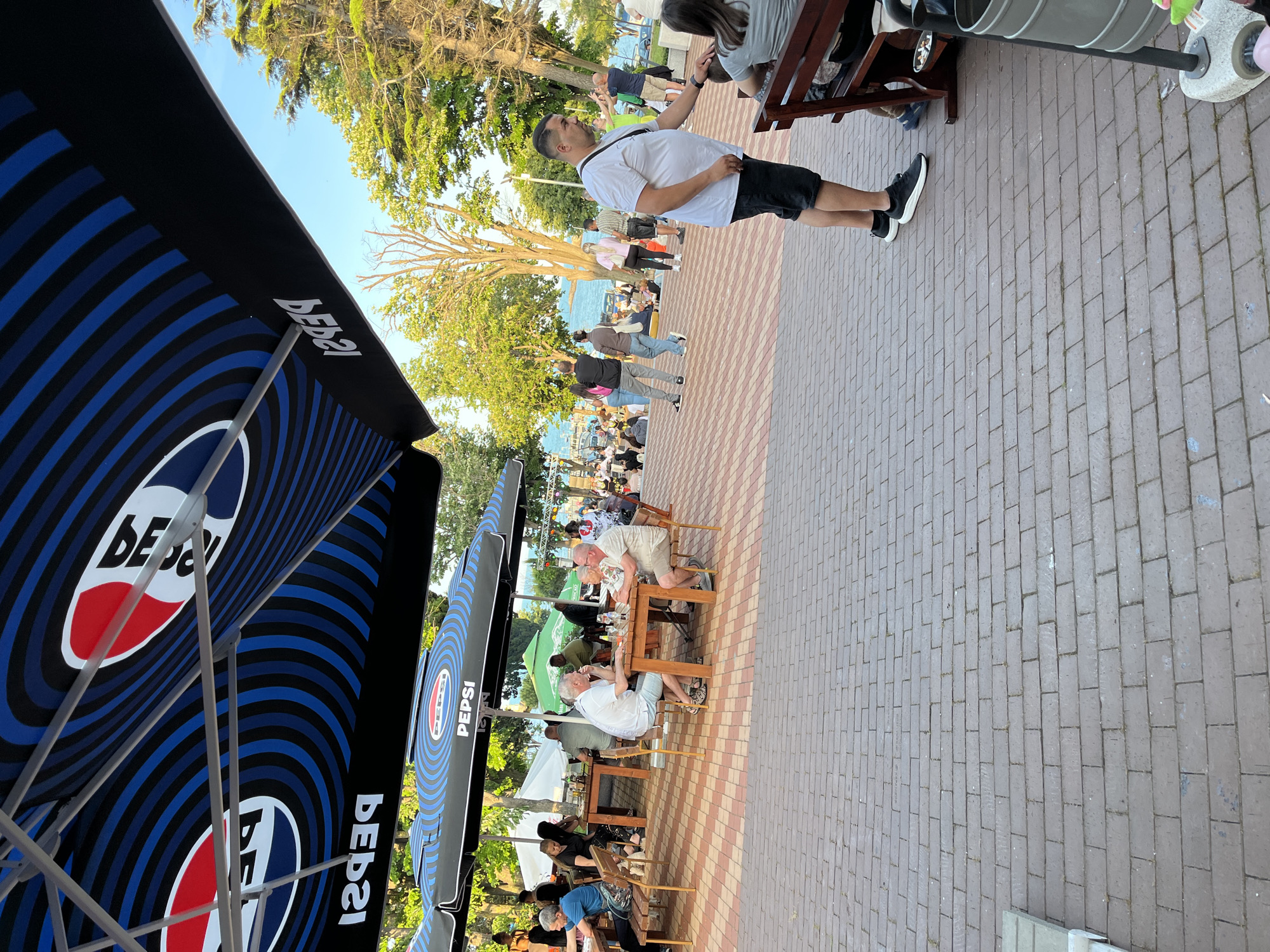 This outdoor scene, presumably in Ahtopol, Bulgaria, depicts a bustling cafe or restaurant area situated alongside a paved promenade with a body of water visible in the background. The sky is clear and blue, indicating daytime.
In the upper portion of the image, multiple large Pepsi-branded umbrellas provide shade. These umbrellas feature black and blue stripes with the iconic red, white, and blue Pepsi logo and text, some appearing inverted due to the viewing angle from below.
The foreground is dominated by a reddish-brown brick pavement. On the far right, a cylindrical grey metal trash can is partially visible. A man wearing a white short-sleeved t-shirt, black shorts, and black sneakers stands in the right foreground, leaning slightly towards a woman whose back is visible as she sits on a wooden bench. His right hand is near her left shoulder.
In the mid-ground, several wooden tables and chairs are arranged under additional Pepsi umbrellas. At one table on the right, two older men are seated, engaged in conversation. One wears a white polo shirt and light-colored shorts with sandals, while the other wears a patterned short-sleeved shirt and light shorts. Glasses are visible on their table. To the left, other patrons are seated at tables, including a woman in a black top.
The background reveals a promenade lined with lush green trees. Beyond the trees and a gathered crowd of people, a calm body of water, likely the Black Sea, is visible. Several people are walking or standing in the mid-to-far background, some near the water's edge. Faintly visible cars suggest a road runs alongside the water. Green and white tents or smaller umbrellas are also present in the background, near the activity by the water.