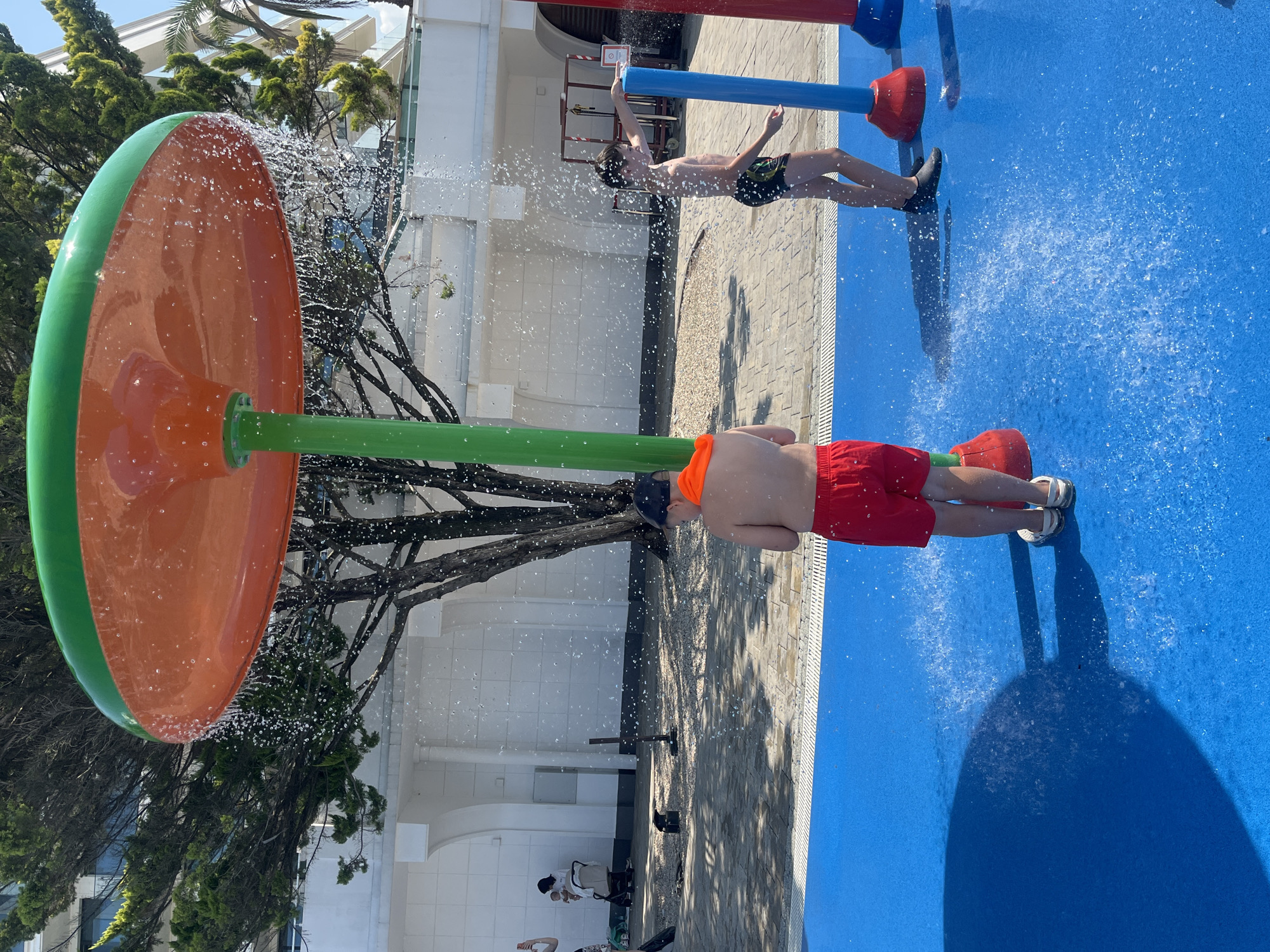 Two young boys are playing in a splash pad, which appears to be located in Sochi, Russia, likely near a resort or hotel given the architecture in the background. One boy, wearing red shorts and an orange towel around his neck, stands under a large, orange and green mushroom-shaped water feature. Water is cascading down from the feature, creating a spray. The other boy, in black and green swim trunks, is standing to the right of the first boy and is also being sprayed by water from a blue pole. The splash pad has a vibrant blue rubberized surface with water jets visible. In the background, there is a white building with arched doorways and trees, including a palm tree, indicating a warm climate. Sunlight is casting shadows on the ground. A few other individuals are visible in the distance, including a woman holding a baby. The overall scene depicts a playful, outdoor recreational activity on a sunny day.