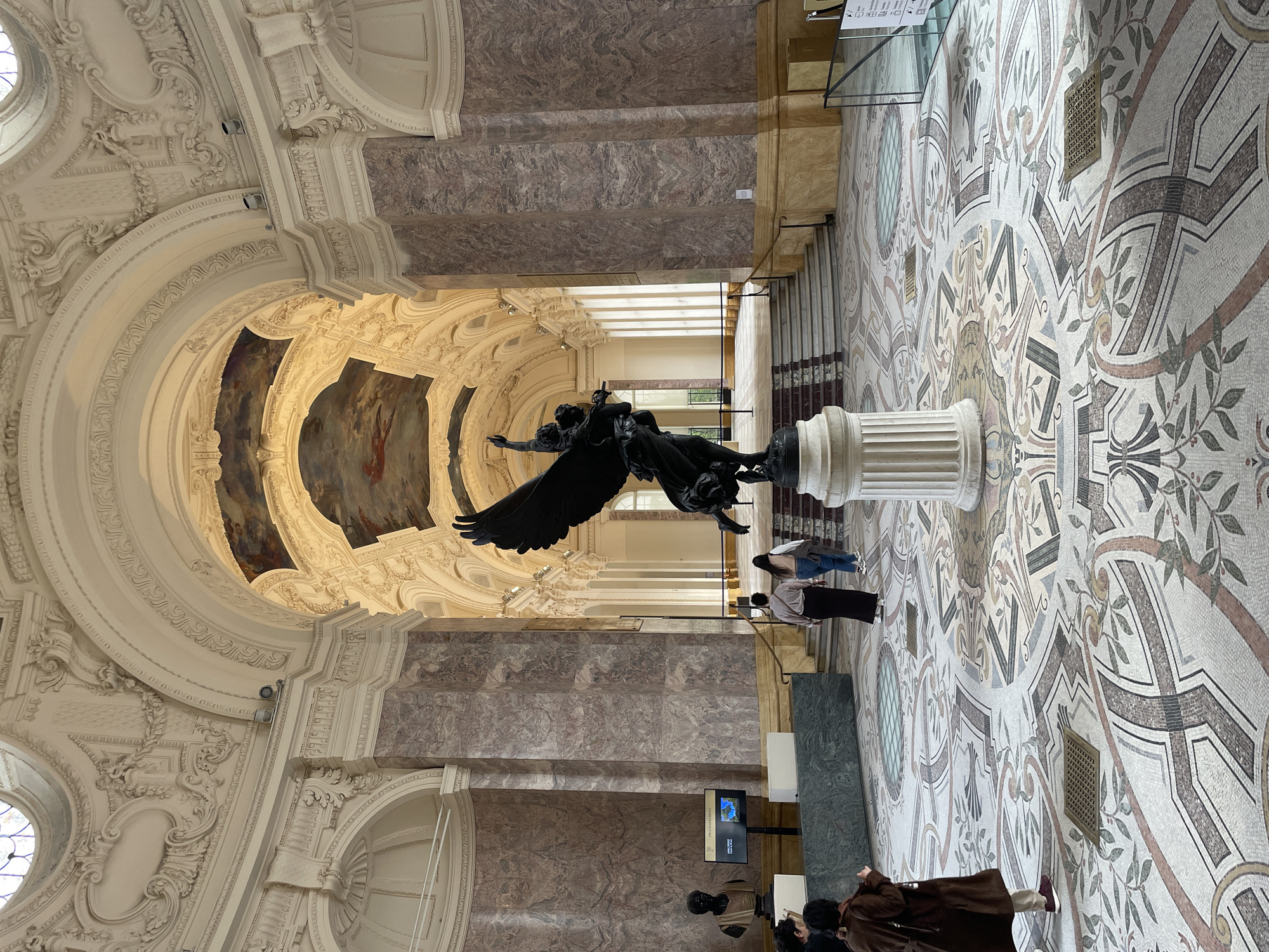 The image depicts a grand interior hall within the Grand Palais, located in Paris, France. At the center foreground, a large, dark bronze statue, identified as "La France portant la Lumière au Monde" (France Bringing Light to the World), stands on a fluted white classical pedestal. The winged female figure is depicted dynamically, with one arm raised, holding what appears to be a torch.
The architectural environment is characterized by ornate classicism. The high, vaulted ceiling is adorned with intricate white stucco relief work and features several large painted frescoes depicting allegorical scenes. Massive, square columns of reddish-brown mottled marble rise from a lighter marble base, supporting the grand structure. The floor is an elaborate mosaic composed of geometric and floral patterns in shades of white, grey, beige, brown, and green. Embedded in the floor are several square metal grates and a circular glass-covered element.
In the mid-ground, a wide, white marble staircase with dark banisters leads to a brightly lit, arched hallway lined with additional columns and arches.
On the left side of the foreground, an information kiosk features a rectangular digital screen displaying "PROCHAINEMENT" (Coming Soon) and a map. Adjacent to this, a dark-colored male bust rests on a white pedestal.
Several individuals are present: In the foreground left, a person in a brown coat and dark trousers stands partially obscured, facing away from the camera. Another person is partially visible behind them. In the mid-ground right, two individuals walk away from the camera, proceeding towards the grand hallway. One wears a light-colored top and dark skirt, the other wears jeans and a light top, carrying a light bag. Low glass barriers and dark metal railings are present in various sections of the hall.