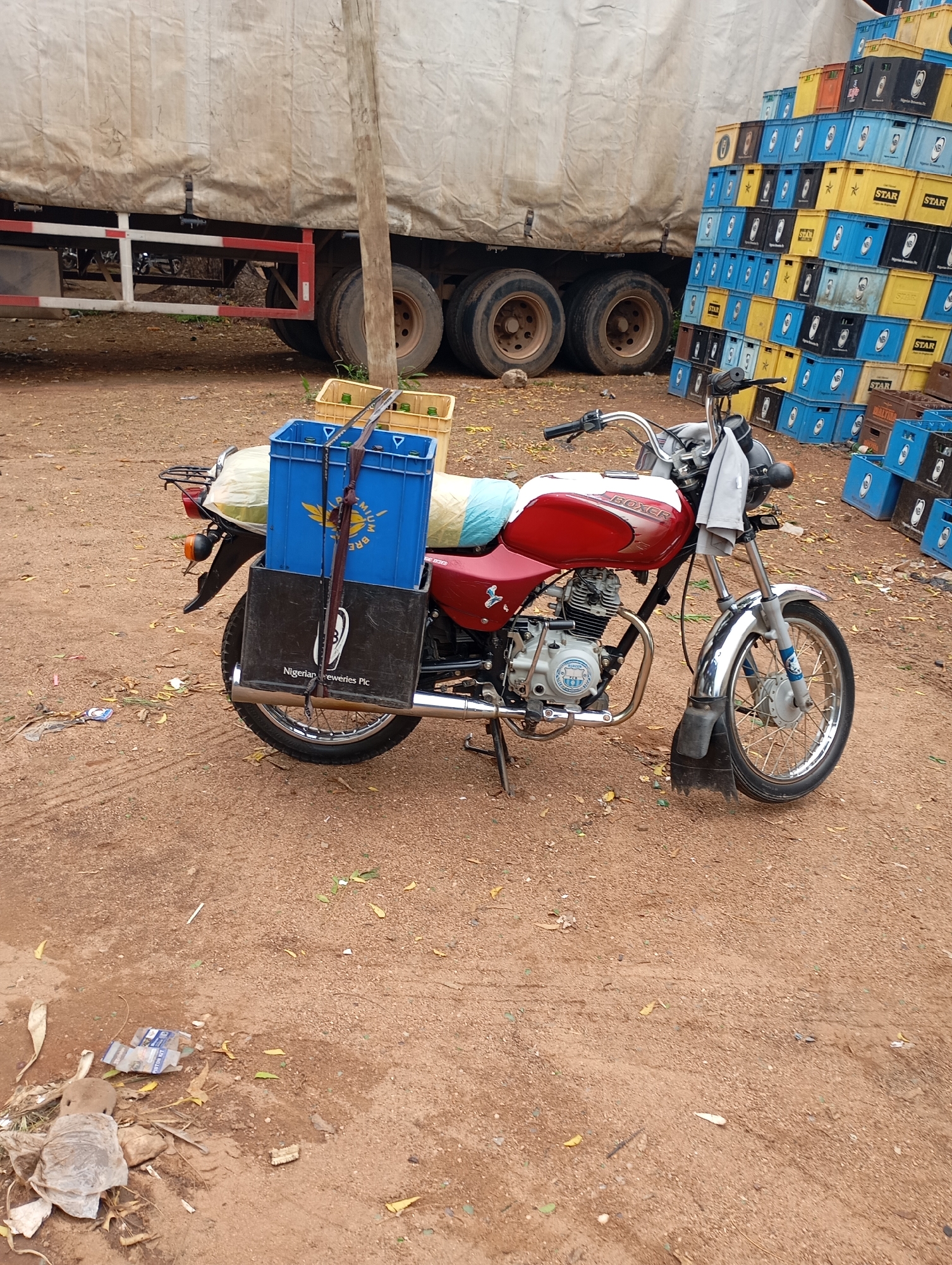 A motorcycle is parked on a dirt surface in front of a large truck. The motorcycle is red and white and has a blue crate strapped to the back, which contains bottles. Next to the motorcycle, there are stacks of beer crates. The truck appears to be a transport truck, with its cover pulled down. The environment suggests an outdoor area, possibly a distribution point or market. The ground is covered in reddish-brown dirt and scattered leaves. The beer crates are various colors, including yellow, blue, and black, and some have branding visible, such as "STAR" and "NB". A wooden pole is visible near the truck. The overall scene depicts a commercial activity involving the transportation and distribution of beverages.