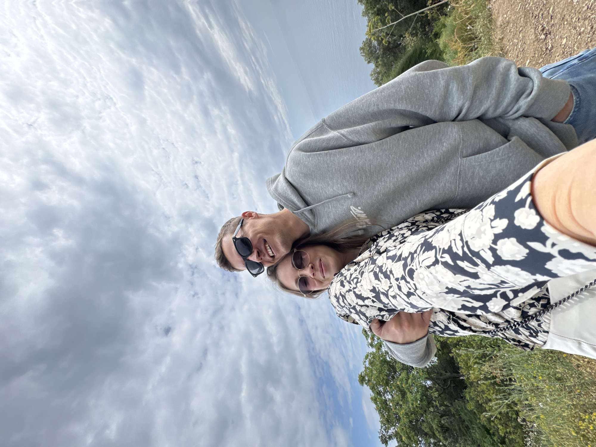 A photograph taken in an outdoor environment, likely Budva, Montenegro, shows a couple. The man, wearing sunglasses and a grey hoodie, is positioned to the left of the woman, who is wearing a floral-patterned top and sunglasses. They are smiling and appear to be embracing or close to each other. The background consists of a cloudy sky, greenery, and some scrub vegetation. The image seems to be taken at a slight angle, possibly as a selfie.