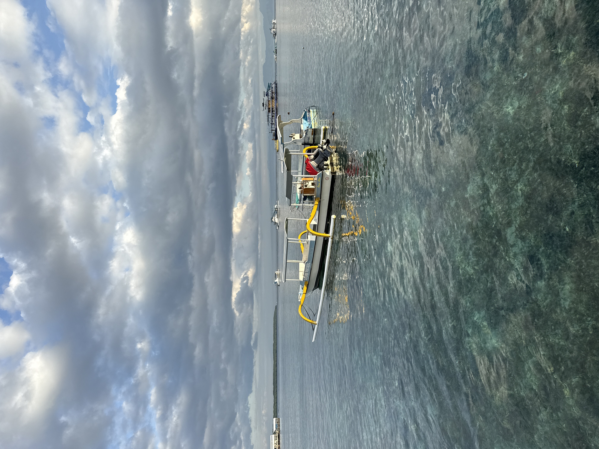 The image shows a motorized boat floating on the water with the sky and clouds visible. The boat is in the foreground, and other boats and land are visible in the distance. The water is clear with a greenish tint, and the sky is cloudy with some blue patches. The location is likely in the coastal region of Nusa Penida, Indonesia, given the types of boats and water conditions.