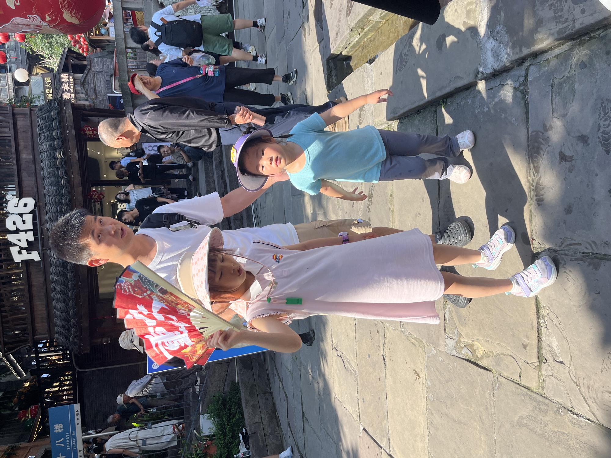 This is a medium shot of two young girls and a man standing on a stone pathway in Yuzhong District, China. The man, positioned in the center-right of the frame, is partially visible from the chest up. He is wearing a white t-shirt and has dark hair. He is holding a red and white folding fan. To his left, a young girl in a pink dress and a wide-brimmed hat is holding the fan. She has dark hair and is looking down. To the man's right, a younger girl in a light blue t-shirt and grey pants is looking towards the camera. She is also wearing a wide-brimmed hat, this one purple. In the background, several other people are walking and standing. Buildings with traditional Chinese architecture are visible. The pathway is made of large, irregular stone slabs. A narrow channel of water runs along the right side of the pathway.