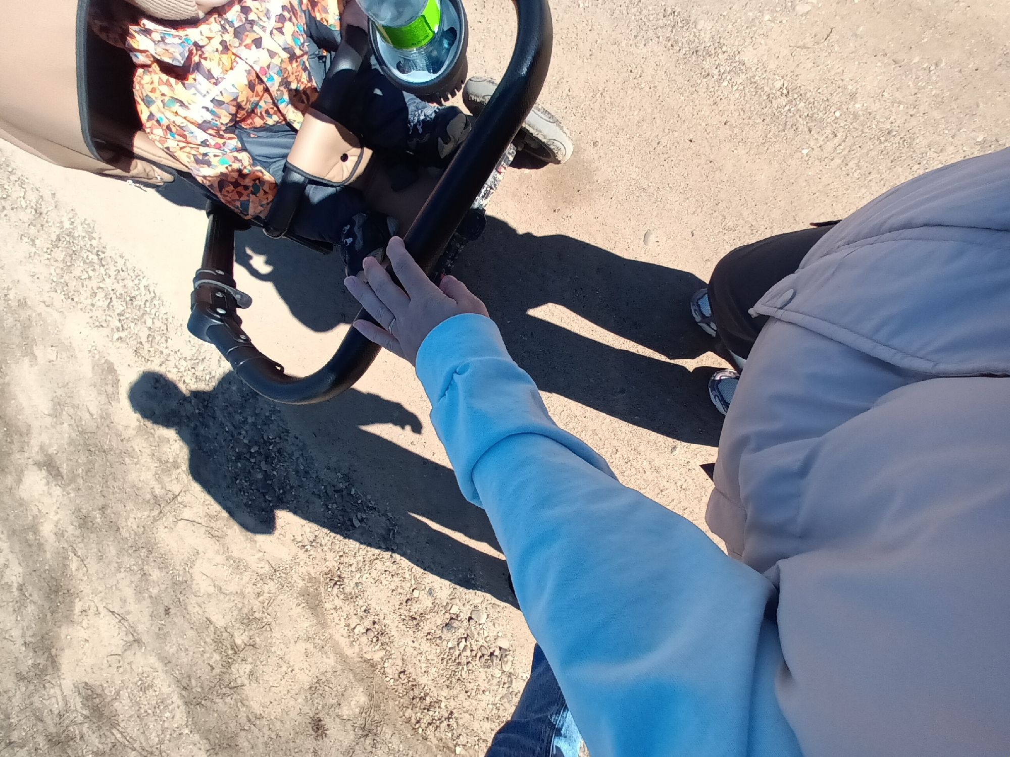The image depicts a top-down view of a person pushing a stroller on a dirt path in Ольховка, Russia. The stroller contains a child. The person pushing the stroller is wearing a light blue sweatshirt and a light-colored jacket. The stroller has a black frame and a brown seat, with a green bottle in a holder. The dirt path has small pebbles and sparse vegetation. The shadows of the person and the stroller are visible on the path.