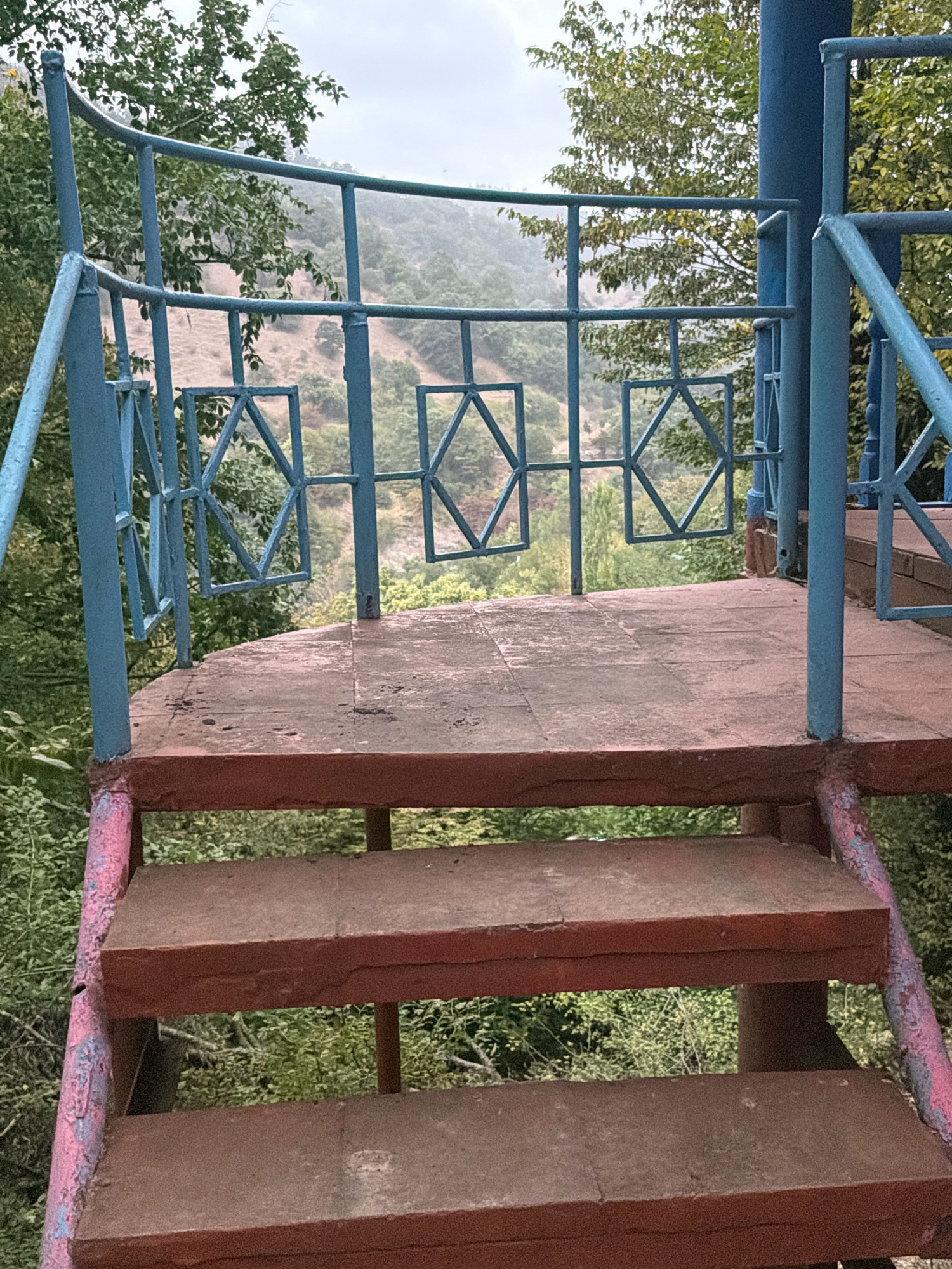 The image depicts an outdoor scene featuring a metal staircase leading up to a circular platform. The staircase and platform railings are painted blue, with some areas of rust and pink paint on the stairs themselves. The platform has a reddish-brown, possibly tiled or concrete surface. Beyond the platform, a lush green, hilly landscape is visible under a cloudy sky. The vegetation in the background appears dense and natural, suggesting a rural or park-like setting. There are no people or animals present in the image. The overall impression is of a somewhat aged or weathered structure in a natural environment.