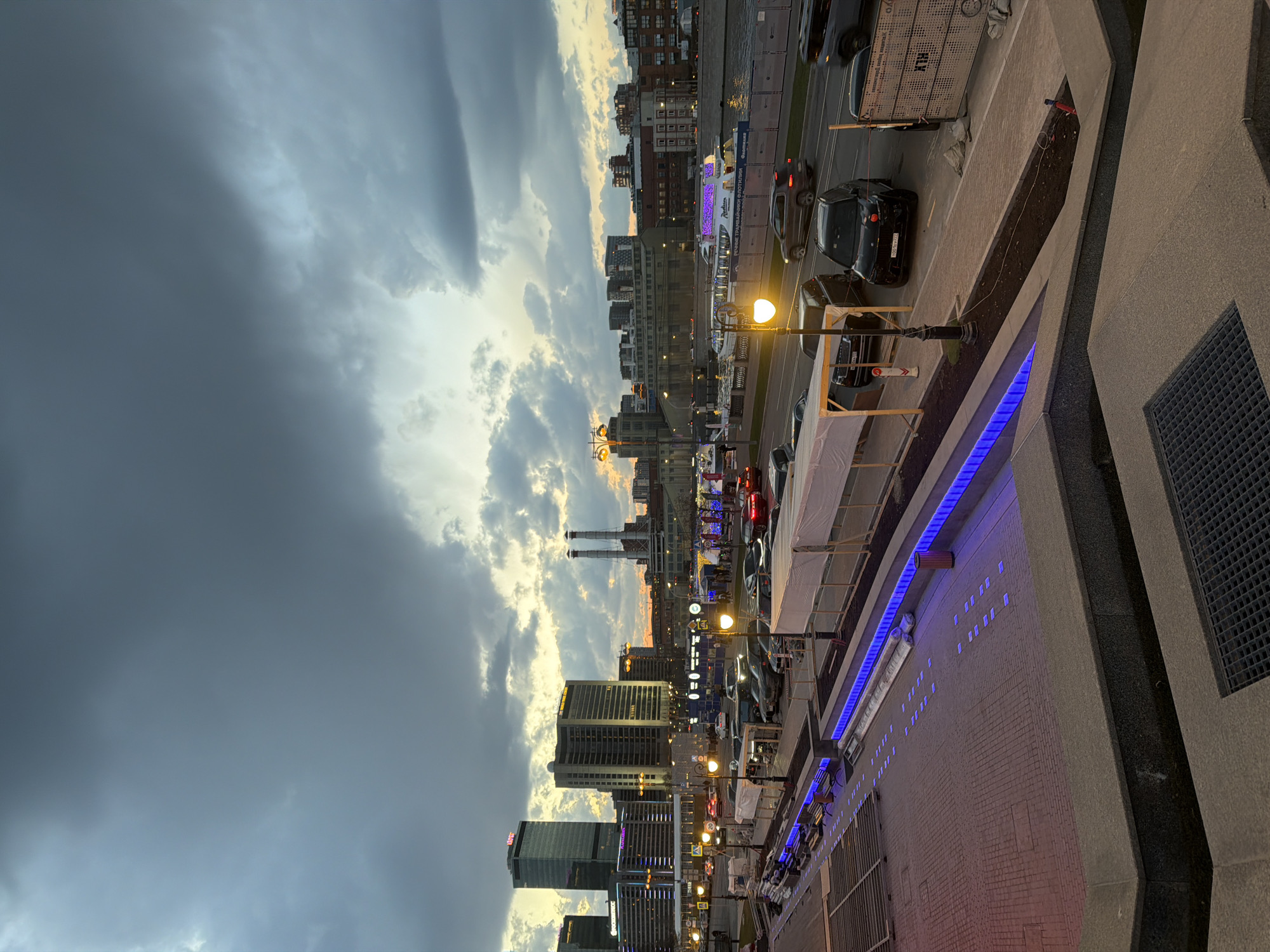 The image depicts a twilight scene in Moscow, Russia, with a view overlooking a cityscape along the Moscow River. Dark, stormy clouds dominate much of the sky, contrasting with patches of lighter, sunset-illuminated clouds near the horizon. The cityscape features a mix of high-rise buildings and industrial structures, including recognizable smokestacks.
A street runs along the riverfront, populated by vehicles and streetlights casting a warm glow. The perspective suggests the photograph was taken from an elevated position, such as a bridge or building, looking down at the urban scene. Features of a building structure are visible at the bottom of the frame, including a brick wall with blue lighting fixtures and a grate.