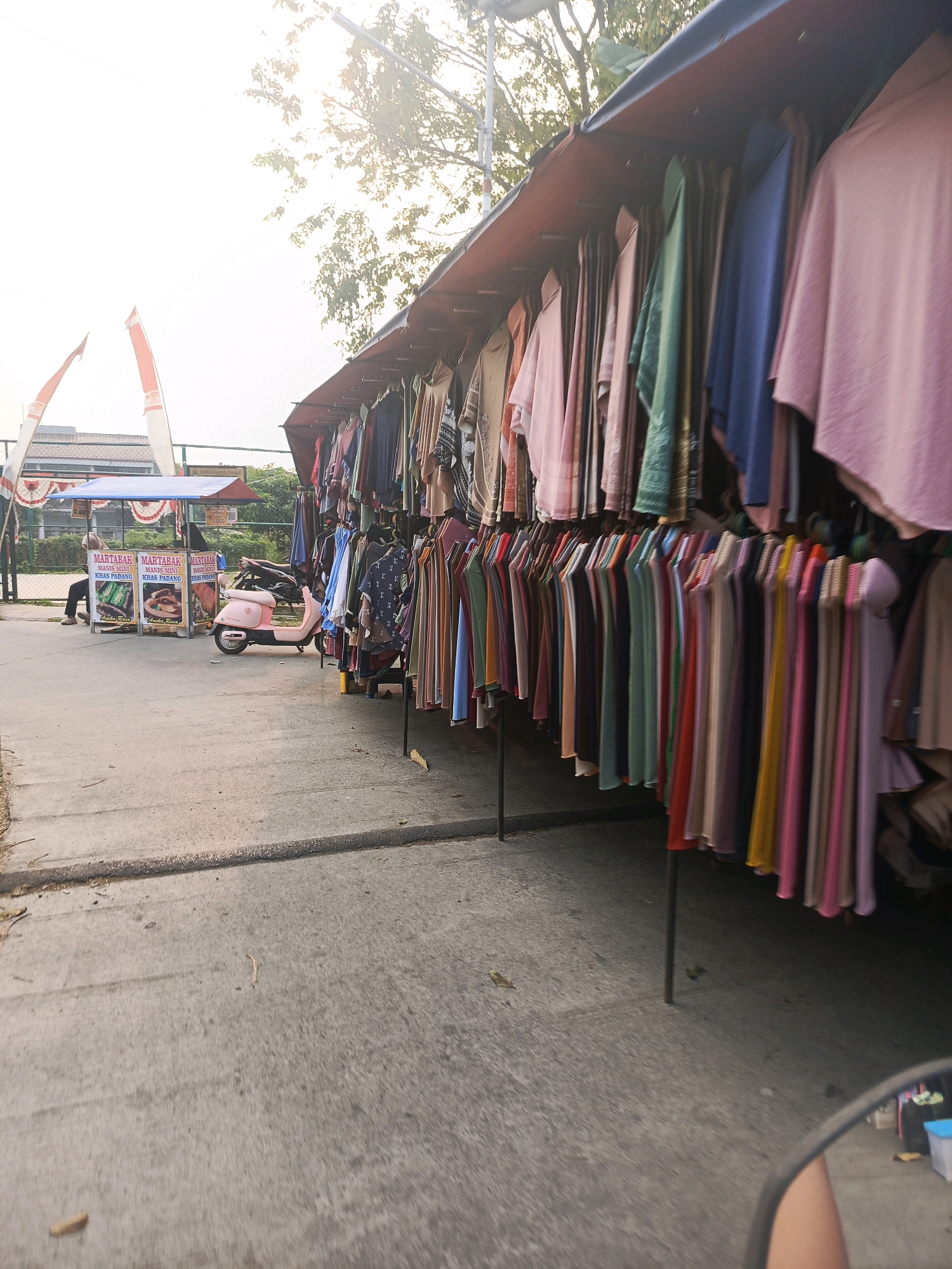In Balaraja, Indonesia, an outdoor clothing market is depicted on a paved street, with stalls set up along the side of the road. A variety of garments, including dresses and blouses in various colors and patterns, are displayed on hangers, creating a vibrant and colorful scene. In the background, a food stall is visible, with advertisements for martabak, a popular Indonesian pancake. A pink scooter is parked near the food stall, suggesting a mode of transportation for vendors or customers. The sky is bright and clear, with sunlight filtering through the trees, casting shadows on the ground. The overall atmosphere is one of lively commerce and daily life in a bustling Indonesian city.