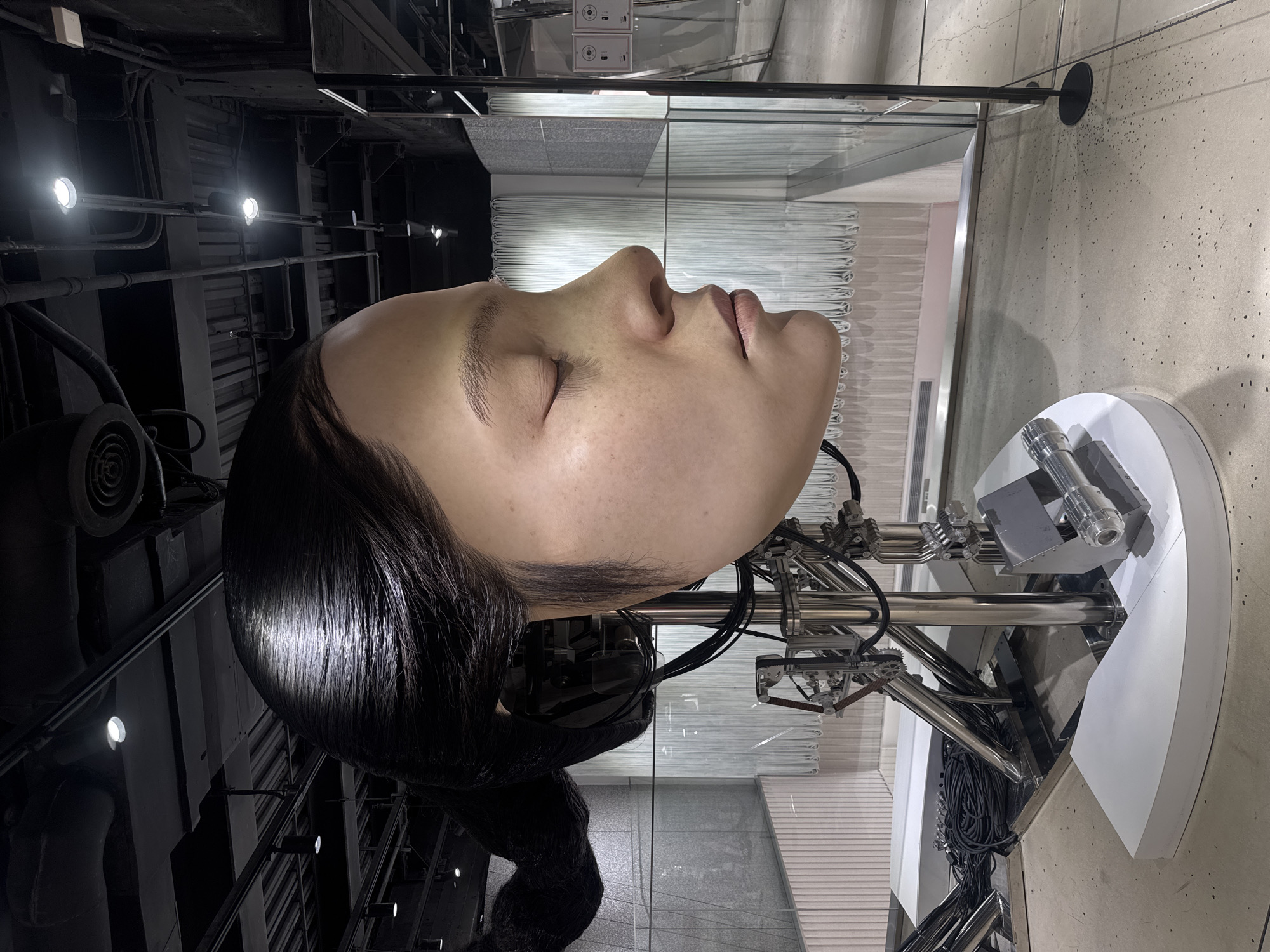 The image shows a large, realistic sculpture of a woman's head and upper neck, with her eyes closed and her head tilted slightly to the right. The sculpture is mounted on a complex, metallic robotic armature that extends down to a white, rounded base. Dark cables and tubes connect to the back of the head and snake down along the armature. The sculpture has dark, shiny hair pulled back into a ponytail. The background consists of a modern interior space with exposed industrial ceilings, ductwork, and lighting fixtures. There are also glass barriers, metallic railings, and light-colored walls with vertical paneling and some translucent curtains. The flooring is a light-colored concrete with dark speckles. This artwork is displayed at the Hyundai Motor Group Innovation Center Shanghai (HMGIC), located in the Huangpu District of Shanghai, China.