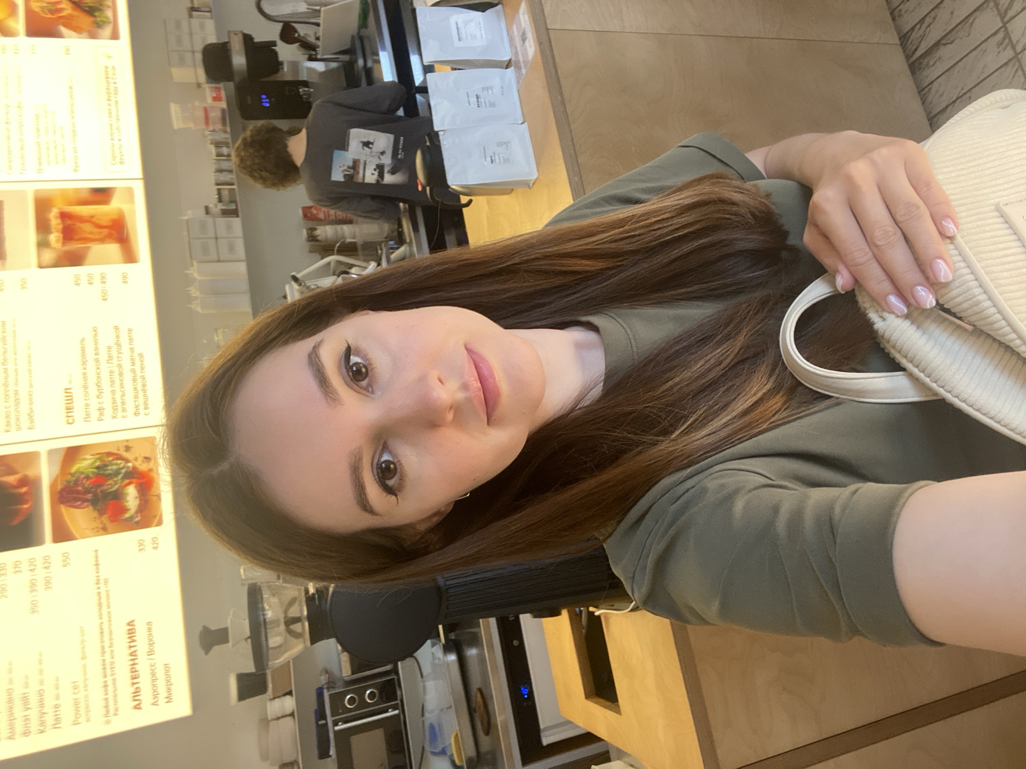 A young woman with long, brown hair and brown eyes takes a selfie in a bright, modern cafe. She is wearing a short-sleeved, olive-green t-shirt and is holding a cream-colored handbag. Her fingernails are manicured with a French tip design. Behind her, a barista with curly hair is visible, wearing a grey t-shirt with a graphic print. In the background, there is a counter with various coffee-making equipment, bags of coffee beans, and a menu board displaying drinks and prices. The menu board, written in Russian, indicates items such as "Американо," "Латте," and "Эспрессо." This scene is likely in Saint Petersburg, Russia, given the Russian text on the menu and the typical cafe aesthetic.