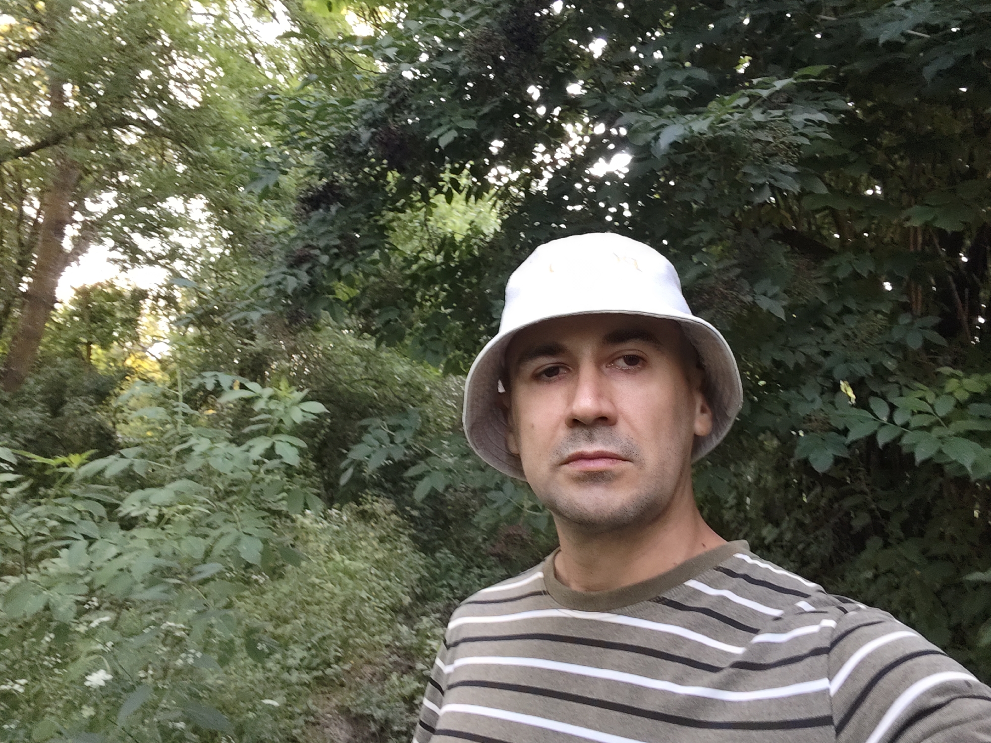 In a medium shot, a man wearing a white bucket hat and a horizontally striped t-shirt is looking directly at the camera. He has short brown hair, a short beard, and light brown eyes. The background is filled with lush green foliage, suggesting an outdoor setting. The lighting appears natural, with some dappled sunlight filtering through the trees, creating a soft glow. The overall scene conveys a casual and relaxed atmosphere. The location is in the city of Akhmetovskaya, Russia.