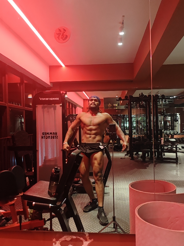 The scene is set in a gymnasium located in the Chongqing High-tech Zone, China, characterized by predominant red ambient lighting. A muscular adult male stands centrally, facing the viewer, posing with his torso exposed and arms flexed slightly outward. He wears black athletic shorts or briefs, black athletic shoes, a blue bandana on his head, and a necklace. His reflection is visible in a large mirror to the right. A small black tripod with a camera or phone mounted on it is positioned directly in front of him, slightly below waist level, suggesting he is being photographed or is photographing himself. To the left, a piece of gym equipment with "HAMMER STRENGTH" text is visible, partially obscured, next to a black padded bench on which a black water bottle rests. The floor is covered with grey diamond-plate textured matting. The ceiling features a prominent linear red LED light strip, three white recessed spotlights, and a circular white plaque with a red Chinese character (福) affixed to it.