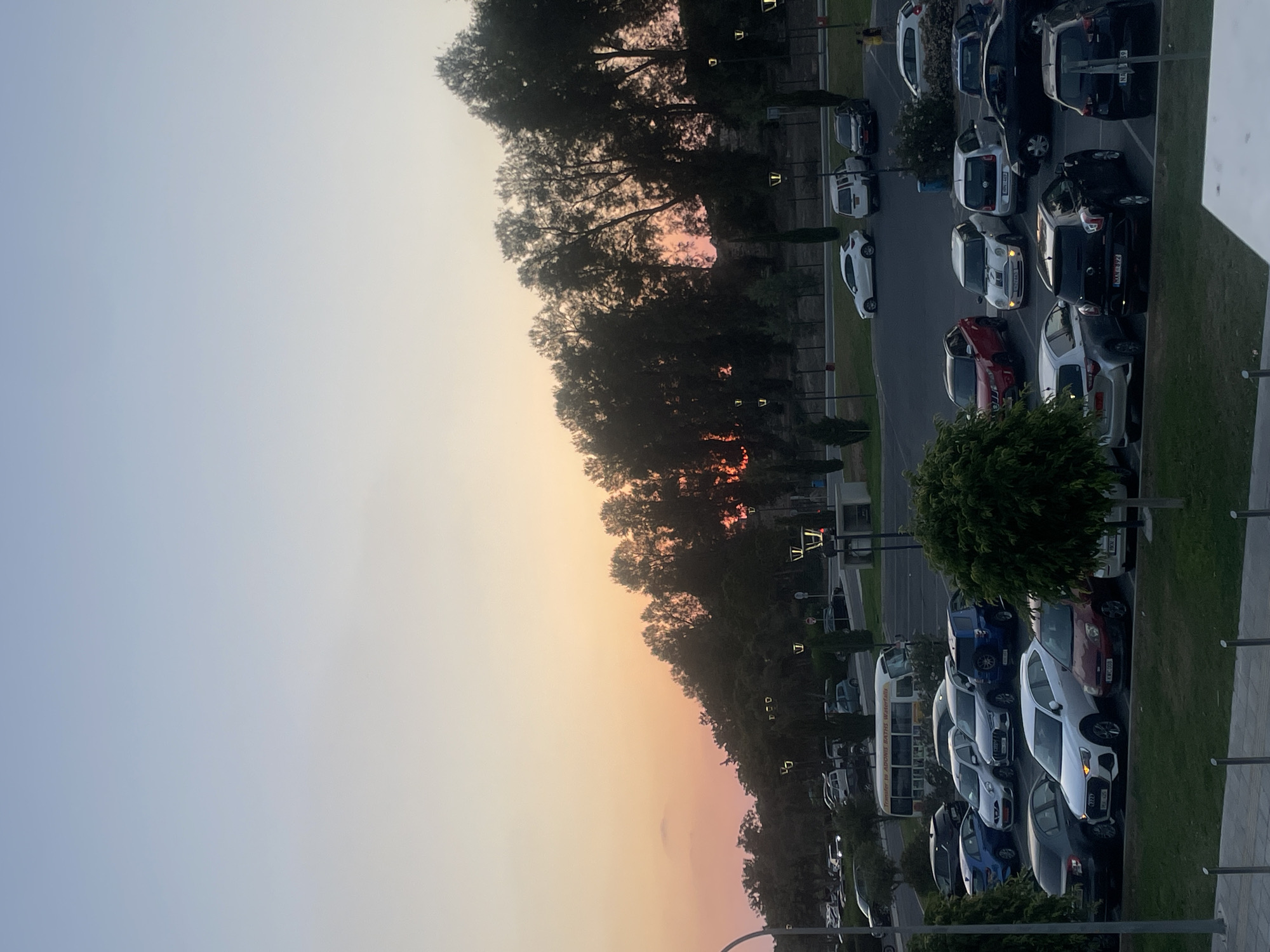 This is a photograph taken at a parking lot near Adonis Baths waterfalls, within the city of Paphos Municipality, Cyprus, during sunset. The sky displays a gradient of soft orange and pink hues, transitioning into pale blue at the top. The foreground is dominated by a parking lot filled with numerous cars of various makes and models, parked in designated spaces. Several streetlights are visible throughout the lot, some of which are illuminated. A small bus with "TRANSFER TO ADONIS BATHS WATERFALLS" written on its side is parked in the background. Lush green trees and foliage line the perimeter of the parking lot, creating a natural backdrop. The overall scene depicts a tranquil evening in a semi-natural environment.
