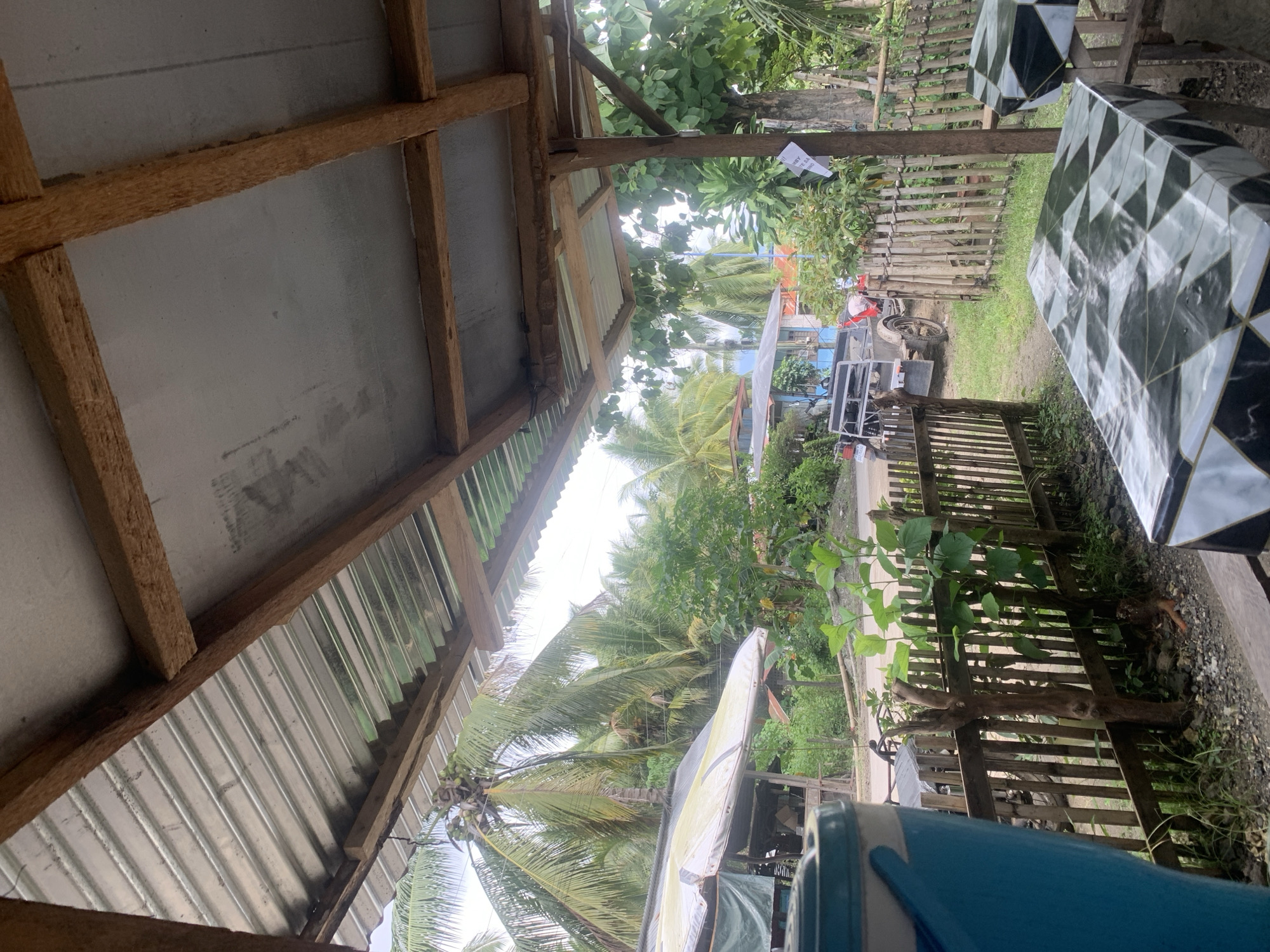 The image captures a view from beneath a corrugated metal roof supported by wooden beams, likely a roadside shelter or eatery in Kiblawan, Philippines. In the foreground, two tables with a black, white, and grey geometric patterned covering are visible. A large blue and white plastic container is partially in the frame on the left. A wooden fence runs horizontally across the mid-ground, separating the immediate area from a street. Beyond the fence, a motorcycle with a sidecar is parked on the road, and further in the distance, a house with a blue roof is visible amidst lush green vegetation and palm trees, characteristic of the tropical climate of Kiblawan. The overall scene suggests a casual, outdoor setting, possibly a rest stop or a small business catering to travelers.