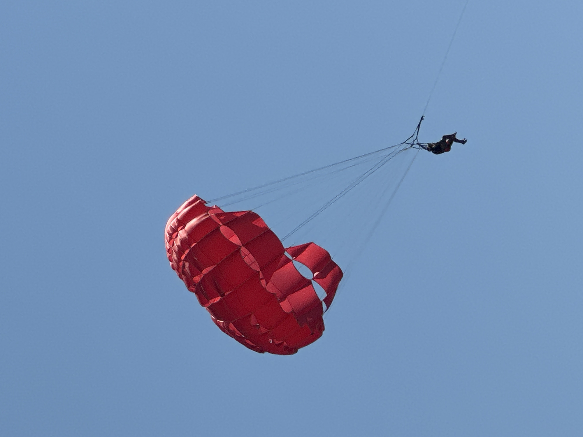 A person is parasailing against a clear blue sky. The parachute is bright red and appears to be inflated and catching the wind. The individual is suspended below the parachute by harnesses and lines, with their legs dangling. The scene is set outdoors with no visible ground or cityscape, suggesting an open area, possibly over water or a scenic landscape. The overall impression is one of aerial activity and recreation.