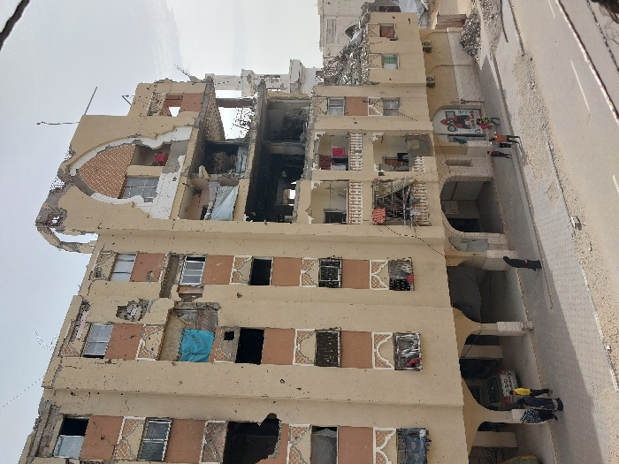 A severely damaged, tall building stands vertically. Significant structural damage is visible, with sections missing and crumbling. Windows are broken, and remnants of furnishings hang from some openings. A road is adjacent to the building. Groups of people are walking along the side of the building. Graffiti decorates the outside of the building. The sky is overcast.