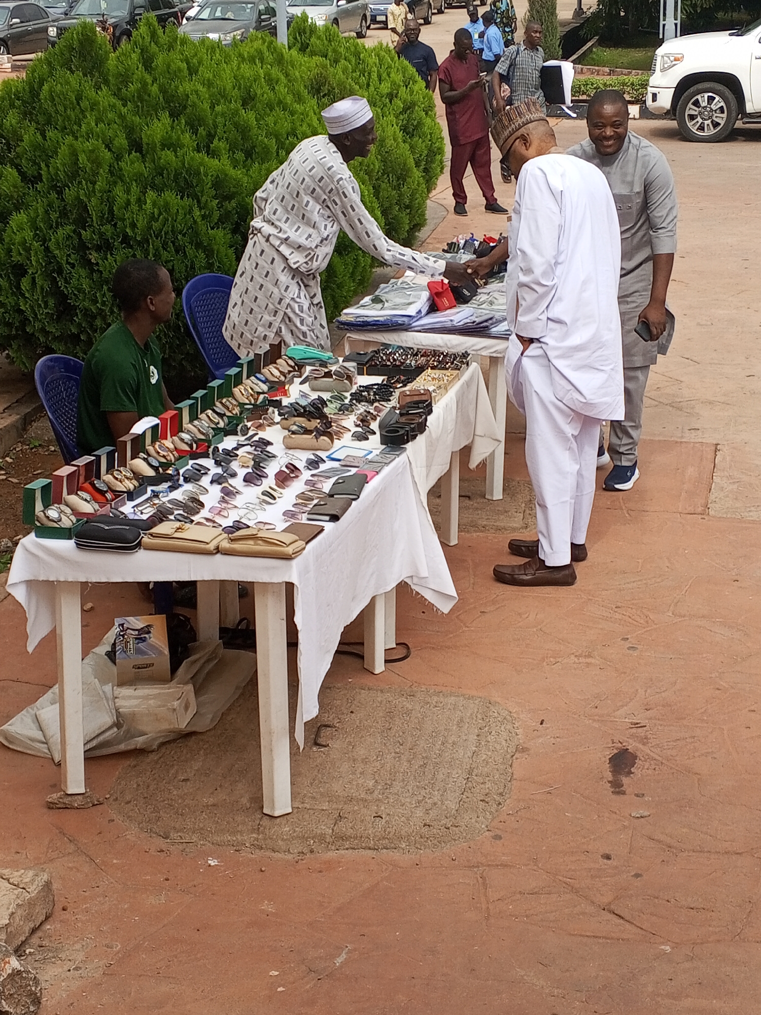 A man is standing behind a table covered with watches, sunglasses, wallets, and purses on a red brick outdoor surface. He is wearing a patterned white robe and a white hat and leaning over the table. Another man is standing in front of the table wearing a white robe. A third man is standing to the right of the second man, wearing a gray robe. A man is sitting behind the table. There is a large green bush in the background, and cars are parked in the background. The location is Jos, Nigeria.