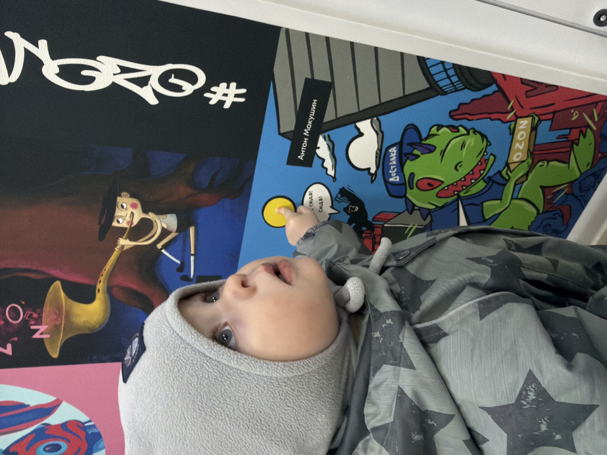 A baby lies on their back, dressed in a warm grey hooded top and a matching grey jacket adorned with dark star patterns. Their head is turned slightly to the left, and their eyes are looking upwards with a curious expression and slightly open mouth. The baby's right hand is reaching out and touching a bright yellow circle, which is part of the colorful graphic surface they are resting on.
The surface beneath the baby is a vibrant tapestry of graphic art, likely a mat, blanket, or wall art. It features various cartoon-like illustrations and text. In the upper left, white stylized text reads "NGRO#" on a black background. Below this, a quirky character with a long nose is depicted playing a golden saxophone.
Further to the right, a blue section displays a black rectangular strip with white Russian text: "Антон Макушин" (Anton Makushin). Nearby, a speech bubble contains the Russian words "сюда! Сюда!" (here! Here!). The most prominent character on the right is a green dinosaur-like creature with red eyes, wearing a blue cap with "ДОСТАВКА" (DOSTAVKA - Delivery) written on it, and a blue shirt. This character is shown holding a brown rectangular object that has "OZON" written vertically, likely referencing the Russian e-commerce company. The scene is indoors, creating a playful and comfortable setting. The Russian text visible on the graphics confirms the location context of Votkinsk, Russia.