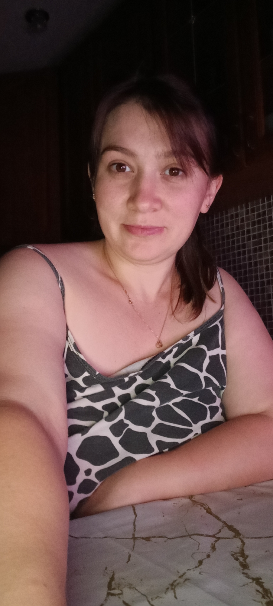 A woman with light skin and brown hair is looking at the camera. She is wearing a gray and white patterned tank top and a thin gold necklace with a small pendant. Her arms are resting on a surface that appears to be covered by a patterned cloth. The background is dark, with some textured tiling visible on the right. There is a light fixture visible in the upper left corner. The setting is indoors, likely a home environment. The specific location in the city of Хохлы, Russia, cannot be determined from the visual information alone.