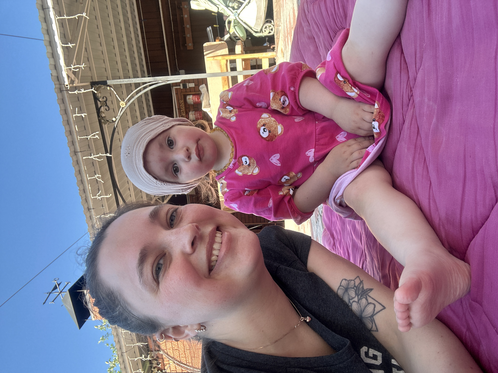 This is a selfie taken outdoors in Yaroslavka, Russia. The subjects are a woman and a young child, possibly her daughter, sitting together on a bright pink, textured surface, likely a blanket or cushion. The woman is in the foreground, looking towards the camera with a wide smile, her head tilted. She is wearing a dark grey t-shirt and has a tattoo of flowers on her left arm. She also has hoop earrings and a delicate necklace with a small pink charm.
The child, positioned behind and slightly to the side of the woman, is looking slightly off-camera. The child is wearing a pink long-sleeved outfit adorned with cartoon teddy bears and hearts, and a light pink, textured bonnet. One of the child's feet is visible, resting on the pink surface.
The background suggests an outdoor setting, possibly a backyard or patio area. There's a hint of a building with a wooden facade and a corrugated roof, along with some decorative string lights overhead. The clear blue sky indicates it is daytime, and the lighting suggests it is sunny, likely late morning or afternoon. The overall mood is joyful and relaxed, captured in the woman's smile and the close proximity of the two subjects. There is no visible text in the image.