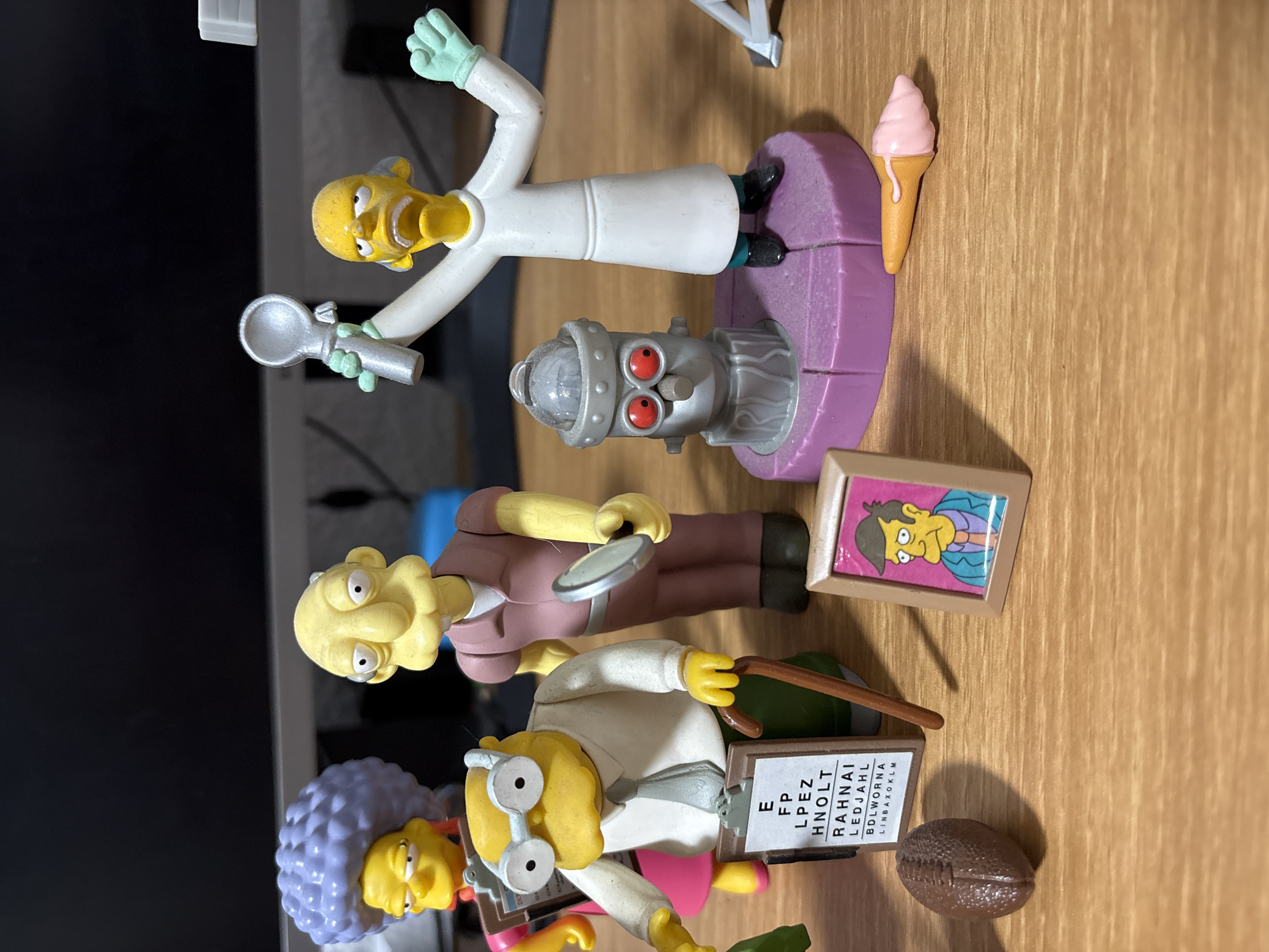 The image depicts a collection of figurines on a wooden surface. The figurines appear to be from "The Simpsons" franchise and include Professor Frink with a machine, a man holding a magnifying glass, and another with a purple hairstyle, glasses, and an eye exam chart. Other objects include an ice cream cone, a framed picture, and a brown oval object. The background includes a black computer screen. There is no indication of the location being Cuernavaca, Mexico.