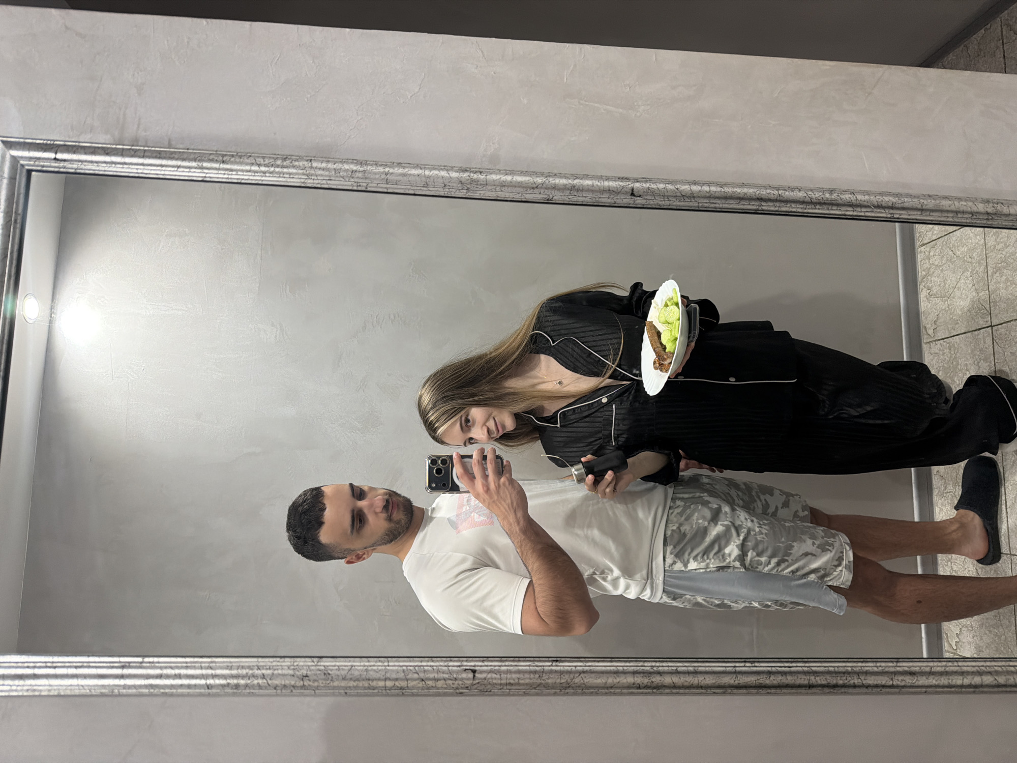 A man and a woman are reflected in a large, ornate silver-framed mirror. The man is holding a cell phone, presumably taking the photo. He is wearing a white t-shirt and camouflage shorts. The woman is wearing black pajamas with white piping and holding a white plate with food on it. They are standing inside a building on a tile floor. The wall behind the mirror is a textured gray. The specific location in Plovdiv, Bulgaria, cannot be determined from this image alone.