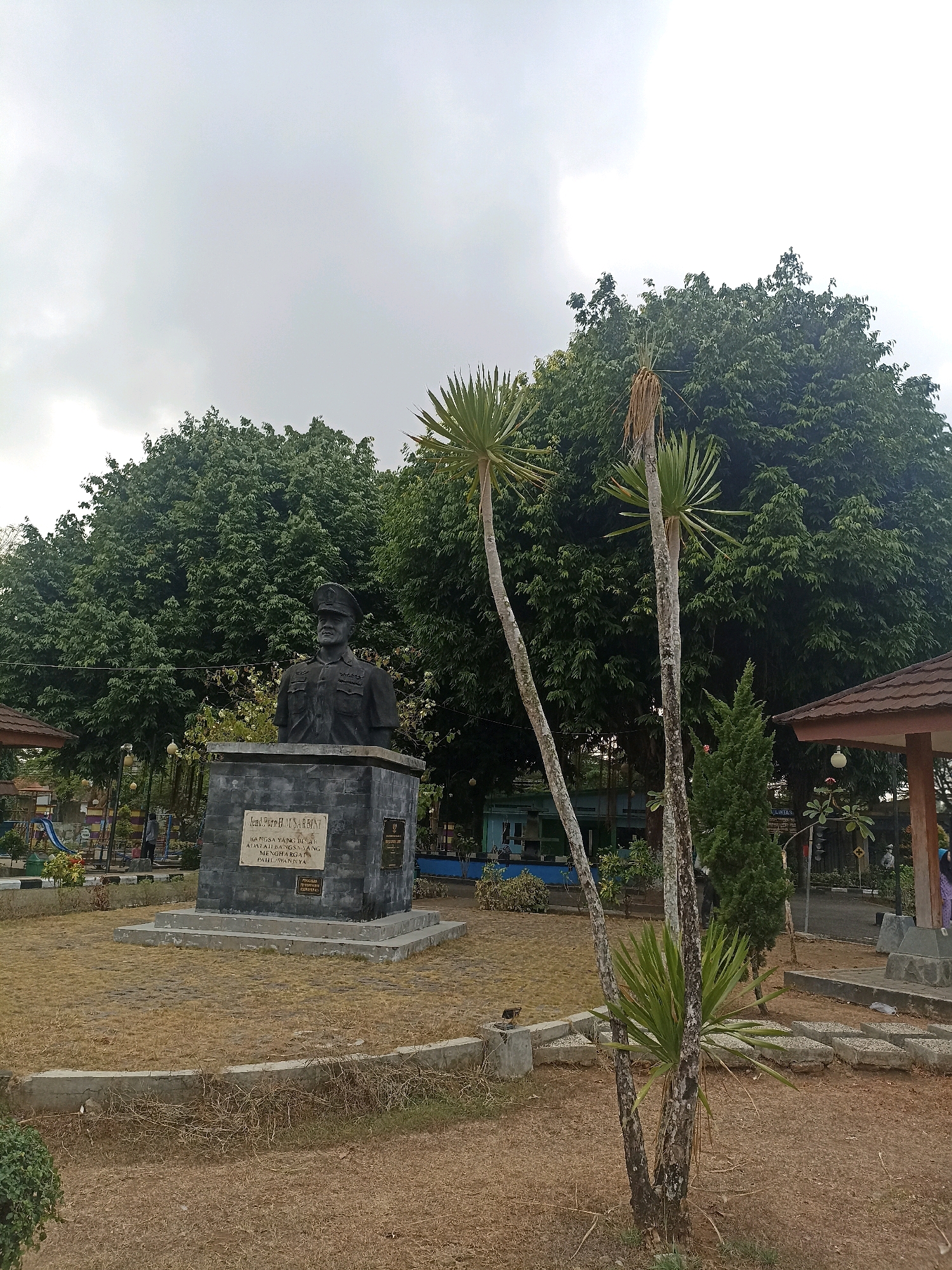 The image captures a bust of a man in military attire, likely a statue, positioned on a stone pedestal. The pedestal has inscribed text, though it is difficult to read clearly. The statue is situated in an outdoor environment with a dry, grassy ground and a backdrop of large, leafy trees. In the foreground, there are two slender palm-like plants. To the left, a playground structure is partially visible, suggesting a public park setting. To the right, a roofed pavilion or gazebo is present. The sky is overcast and appears to be gray. The location is identified as Panjer, Indonesia.