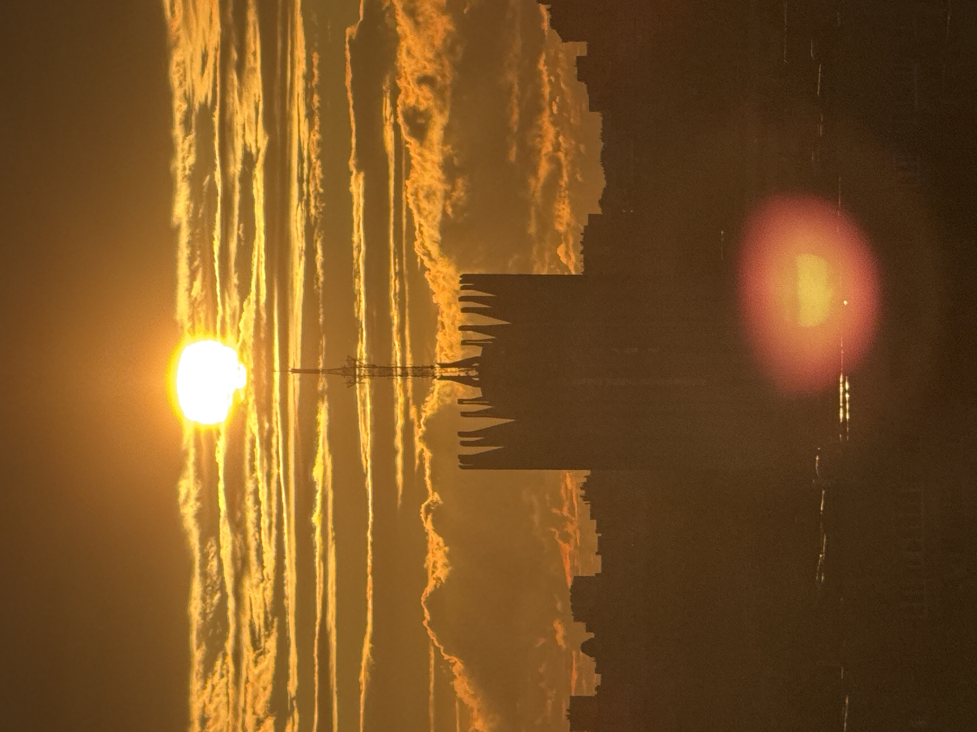 The image captures a sunset over a city skyline, likely in Russia given the context. The sun, a bright, almost white orb, is positioned in the upper center of the frame, partially obscured by a dense layer of horizontally striated clouds. These clouds are illuminated by the sun, creating a dramatic golden and orange gradient across the sky.
Dominating the mid-ground is the silhouette of a tall, distinctive building with a crenellated top, reminiscent of a fortress or a castle. A slender antenna or spire extends vertically from the apex of this structure, piercing the cloud layer. To the left and right of this central building, the outlines of other urban structures and buildings are visible, appearing as dark, indistinct shapes against the vibrant sky.
In the foreground, the lower portion of the image shows a hazy, diffused reddish-orange circle, which appears to be a lens flare or a reflection of the sun. Below this, further indistinct shapes suggest the presence of more buildings and possibly distant lights, all rendered in shadow and silhouette against the warm tones of the sunset. The overall atmosphere is one of dramatic natural light interacting with the urban landscape.