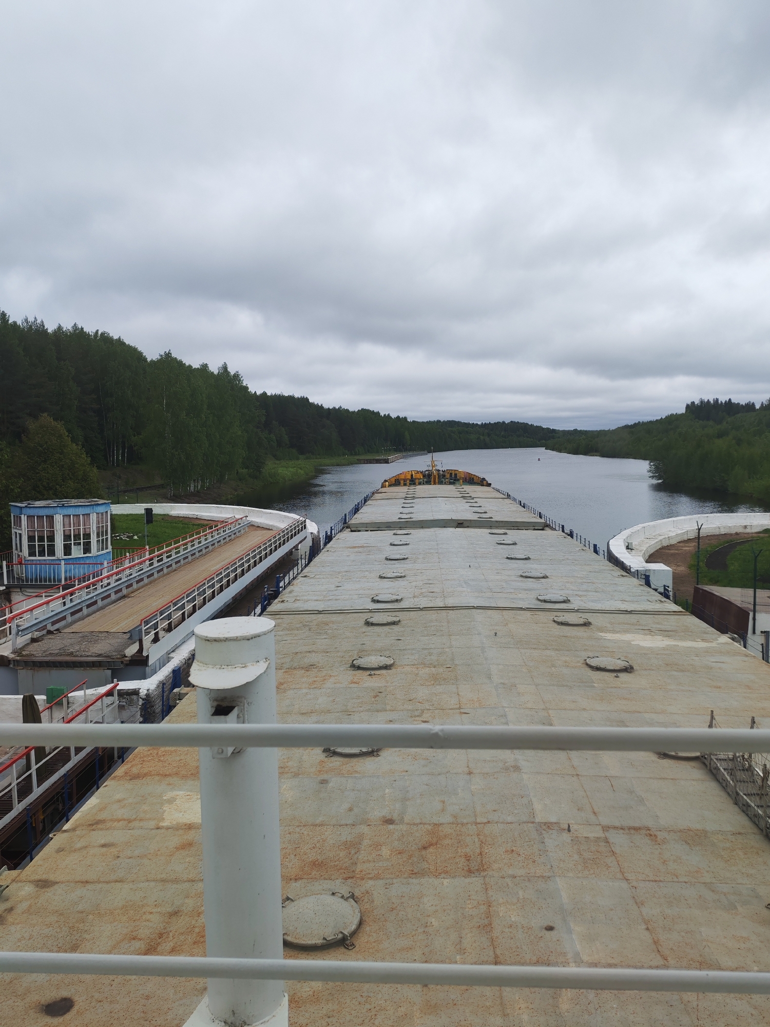 A high-angle, wide-shot photograph depicts the deck of a barge or ship situated on a river or canal, likely in Alekseevskoe, Russia. The foreground shows a large, flat, gray deck with several circular hatches. A white railing runs across the bottom of the frame, and a white cylindrical post is visible.
To the left, a control station with a blue roof and white trim is connected to the ship via a walkway with red railings. Beyond the ship and the station, the river flows between forested banks under an overcast sky. The river appears calm, with a few ripples on the surface. The trees along the banks are lush and green. The sky is uniformly gray with low-hanging clouds.