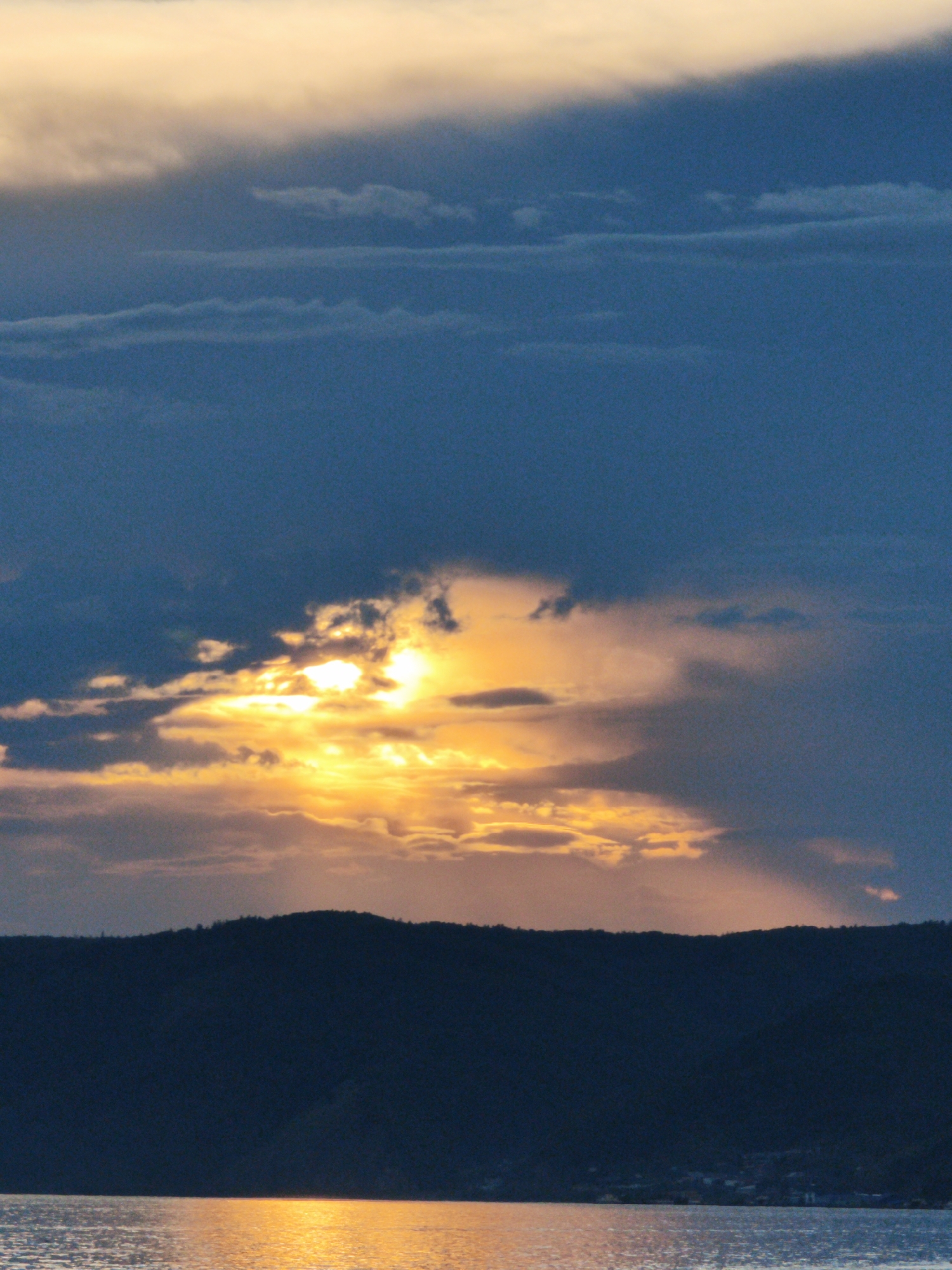 The image depicts a dramatic sunset over a dark, silhouetted mountain range, likely overlooking a body of water. The sky is filled with a gradient of colors, transitioning from deep blues at the top to vibrant oranges and yellows near the horizon, where the sun is partially obscured by clouds. The water in the foreground reflects the warm hues of the sunset, creating a shimmering effect. The mountain range, which appears to be covered in trees, forms a dark, undulating line across the middle ground. There are no visible people or specific structures that would immediately identify the location. However, the description of Listvyanka, Russia, suggests this scene could be a view of Lake Baikal from the shore.