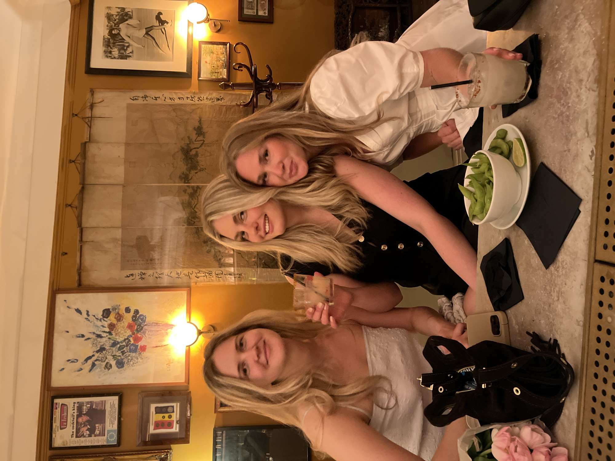 Three young Caucasian women are seated at a bar, posing for a photo. The woman closest to the camera is wearing a white top. To her right is a black handbag and a bouquet of pink flowers. The woman in the middle is wearing a black dress with gold buttons. The woman in the background is wearing a white blouse. On the bar are a cocktail, a bowl of edamame, and two black napkins. The wall behind them is decorated with multiple framed pictures and a light fixture. The location appears to be a restaurant or bar, potentially in London, United Kingdom, based on the style and ambiance.