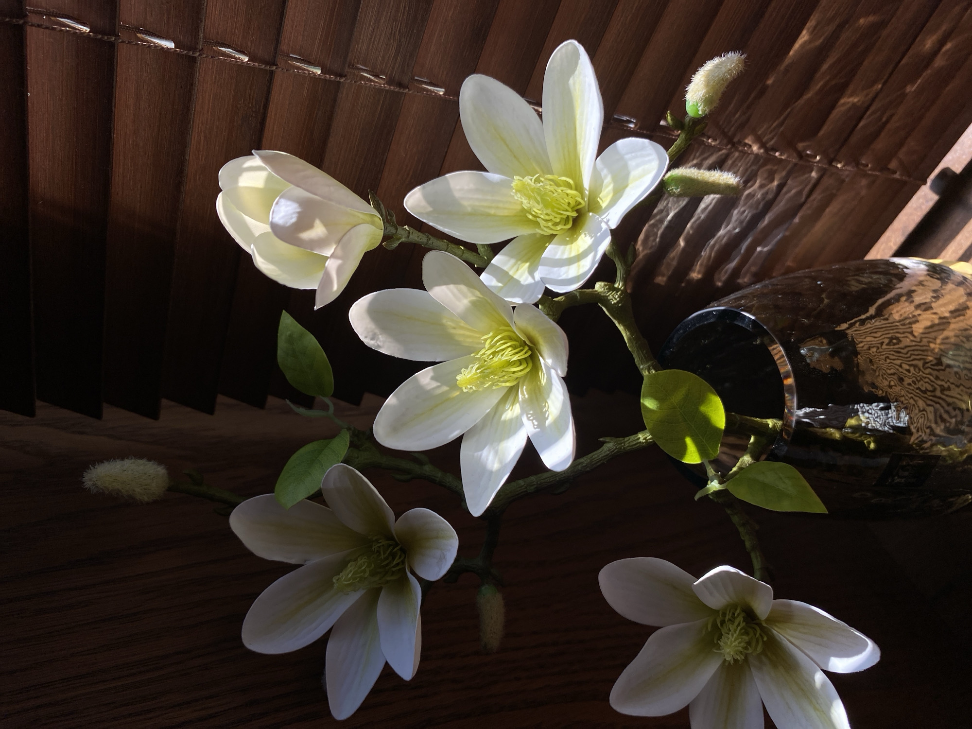 The image depicts a floral arrangement, likely artificial, featuring white magnolia-like blossoms with yellow centers and budding sprigs. The arrangement is positioned on a dark wooden surface in front of a blurred background with dark brown vertical blinds and an abstract glass vase. The scene is illuminated with strong directional light, casting shadows across the wooden surface. There are no people visible in the image. The scene is unlikely located in Votkinsk, Russia, as this is not evident from the image.