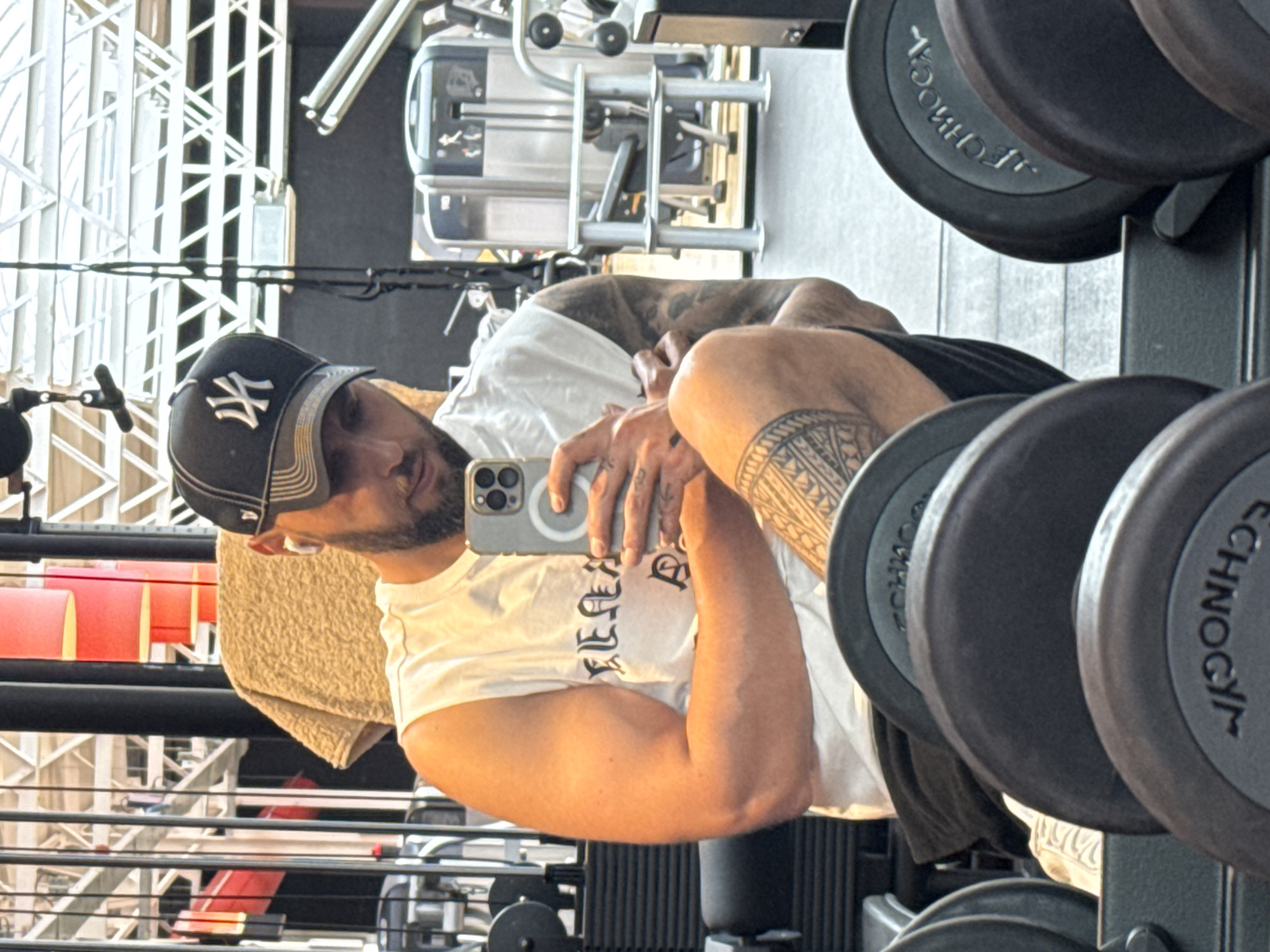 A man with a beard and a black baseball cap with a white logo is taking a mirror selfie in a gym. He is wearing a white sleeveless shirt and black shorts. He has tattoos on his right arm and leg. In front of him, there are several dumbbells. In the background, gym equipment is visible. The image appears to have been taken inside a gym in Emmen, Netherlands.
