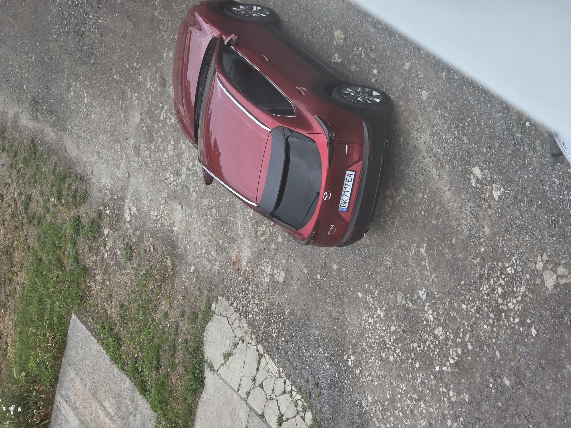 A dark red Mazda CX-30 is parked on a gravel surface next to a concrete path with grass growing between the cracked slabs. The vehicle's license plate reads "BK 7117 EA". The scene appears to be in Dubno, Ukraine. The environment is outdoors with a slightly overcast sky creating soft lighting. No people are visible in the image.