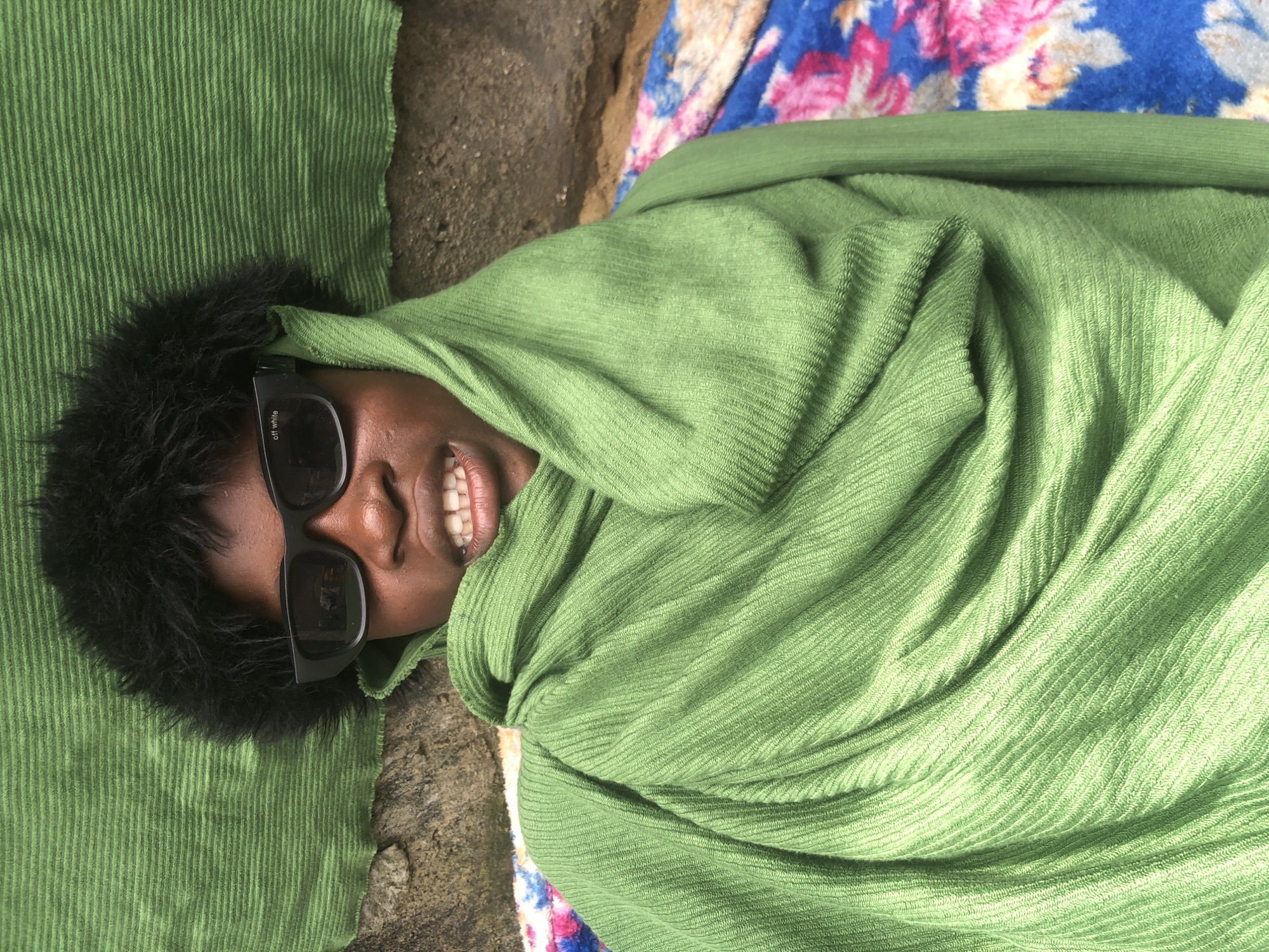 A close-up, medium-shot photograph shows a person lying down, wrapped in a green scarf. The person is wearing black sunglasses with the text "off white" printed on the left lens. Their mouth is open, revealing teeth. The background consists of a textured green fabric, a stone wall, and a colorful blanket. The scene appears to be outdoors, possibly in a residential area of Ado, Nigeria.