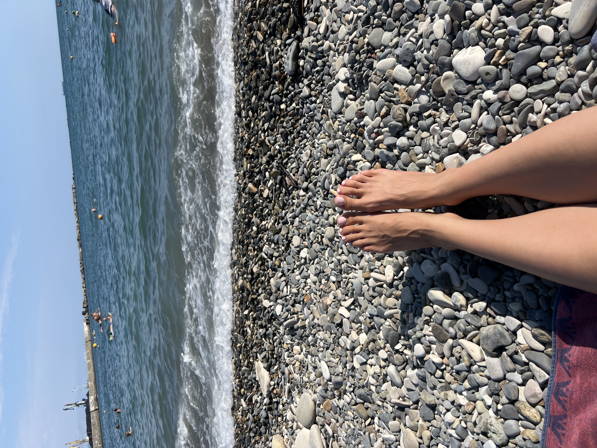 This is an eye-level, outdoor, close-up shot of a person's bare feet resting on a pebble beach. The feet are tanned and have light pink nail polish. The pebble beach is composed of various sizes and colors of smooth, rounded stones, with some larger stones interspersed. The sea is visible in the background, with gentle waves breaking on the shore. The water is a deep blue and appears to be a calm sea. There are several people in the water, some appearing to be swimming or floating on inflatable devices. In the distance, a breakwater or pier with industrial cranes is visible, along with buoys and a small ship. The sky is a clear blue. This scene is consistent with a beach location in Tuapse, Russia, a city on the Black Sea coast known for its pebble beaches and port.