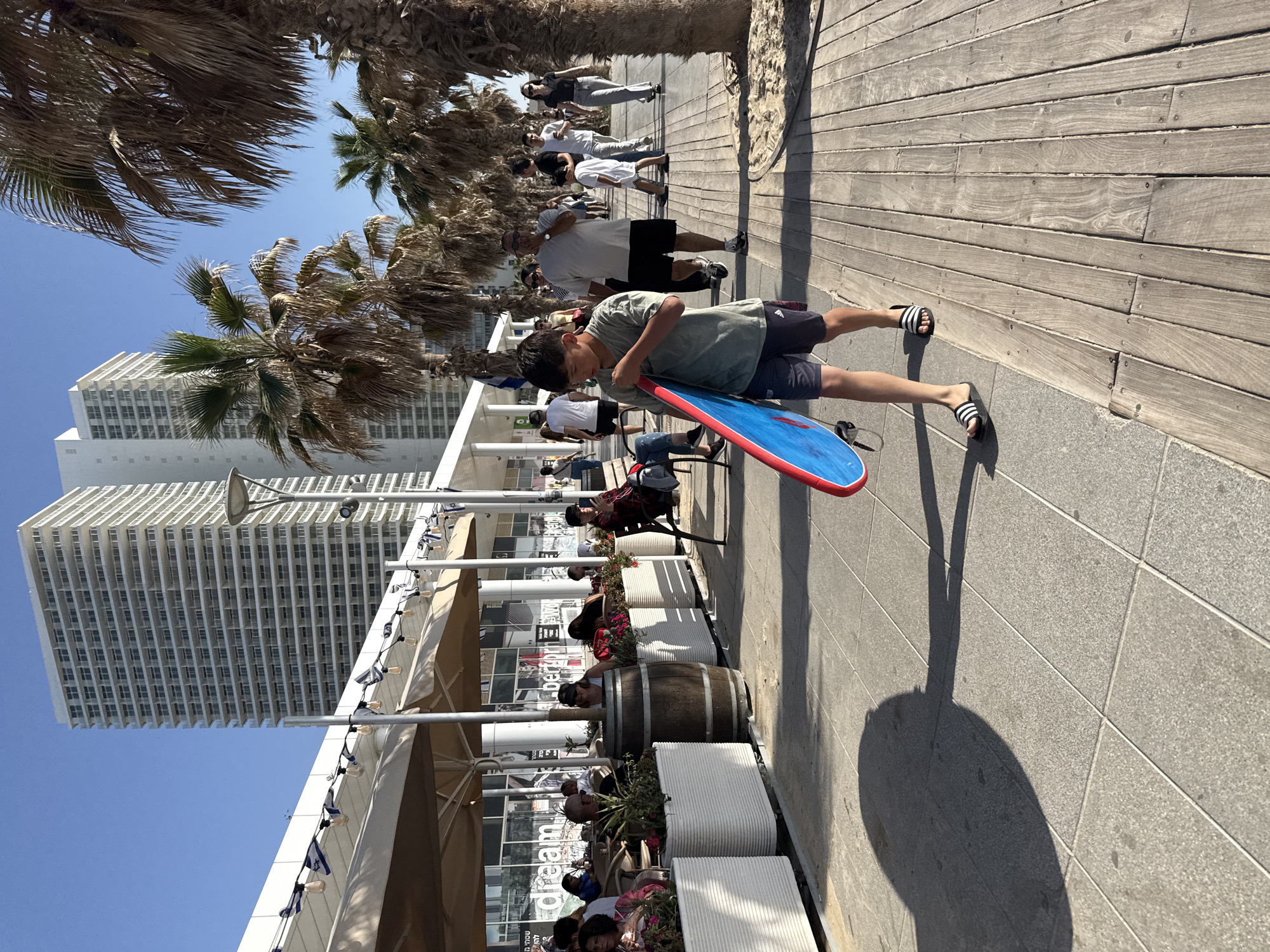 A medium shot depicts a boardwalk scene in Bat Yam, Israel, featuring a young boy carrying a blue and red boogie board. He wears a green t-shirt, dark shorts, and striped sandals, and is walking along a concrete path adjacent to a wooden boardwalk. A group of people is strolling behind him on the boardwalk, and a line of palm trees runs parallel to the walkway. A building with a white facade, possibly a hotel or apartment complex, rises in the background. A restaurant with outdoor seating is visible, featuring white planters, wooden barrels, and umbrellas. The lighting suggests it is daytime, casting shadows from the palm trees and objects along the path.