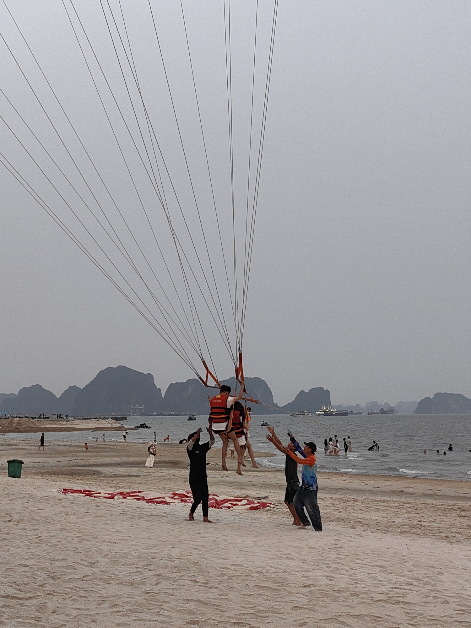 The image shows a beach scene in Bai Chay Ward, Vietnam. In the foreground, two people are being prepared for parasailing. Three men on the ground are managing the activity. One man is in black clothing, and the other two are wearing casual clothing. Scattered red and white parachutes are on the sand. People are swimming and enjoying the beach in the background, and boats are on the water near some islands. The sky is overcast, and there's a pier on the left side of the image.