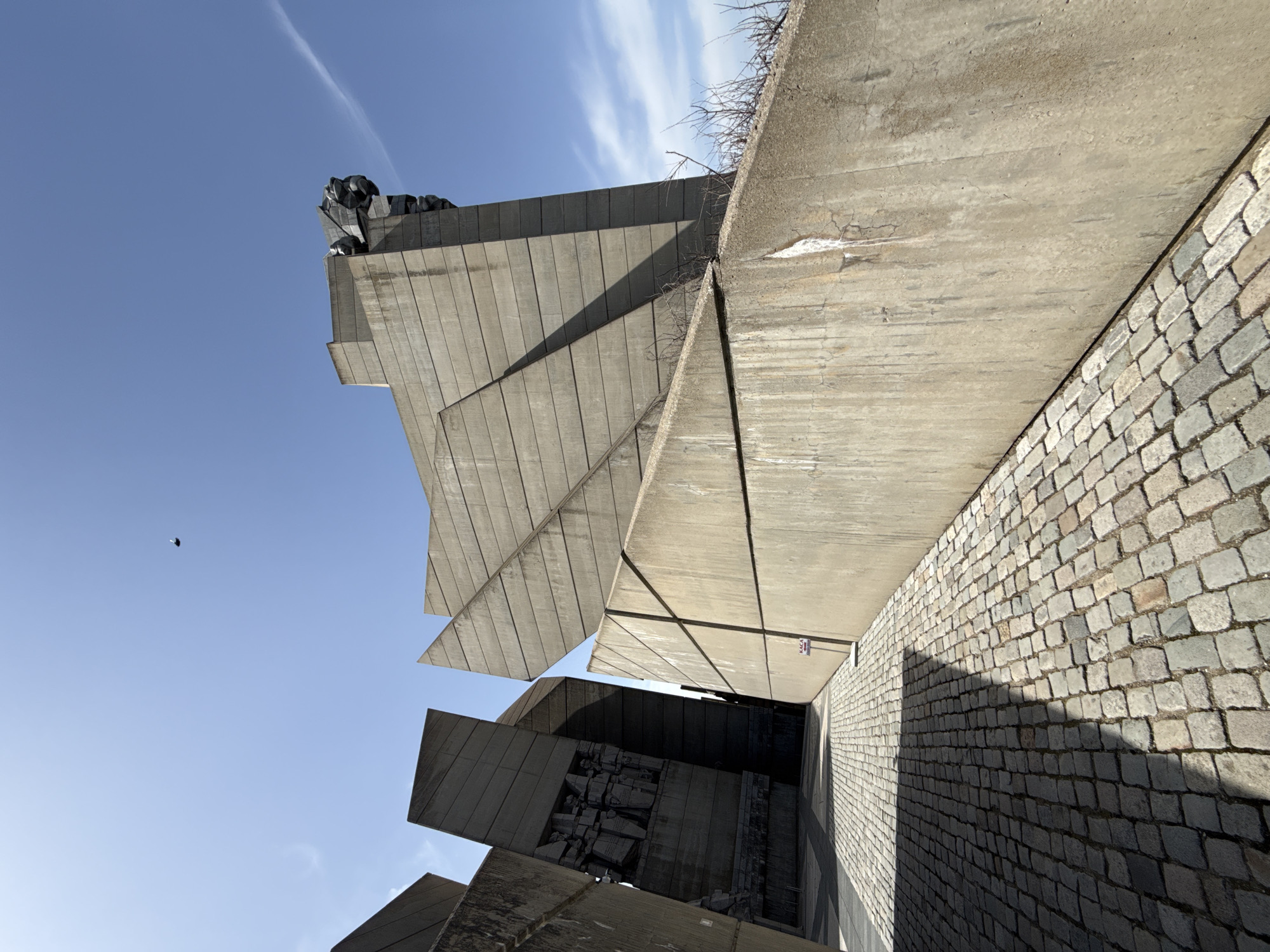 The image shows a monumental concrete structure against a bright blue sky, likely the Founders of the Bulgarian State Monument in Shumen, Bulgaria. The structure is composed of large, angular concrete slabs, some smooth and others with vertical ridges. The slabs intersect and overlap, creating a complex geometric form. On top of the structure, a dark, indistinct sculpture can be seen. The foreground features a cobblestone pavement that slopes downwards and to the right. The perspective is from a low angle, emphasizing the monument's scale and imposing presence. A small, dark bird is visible against the sky. Bare branches sprout from the top edge of the concrete. A small sticker is attached to the concrete wall.