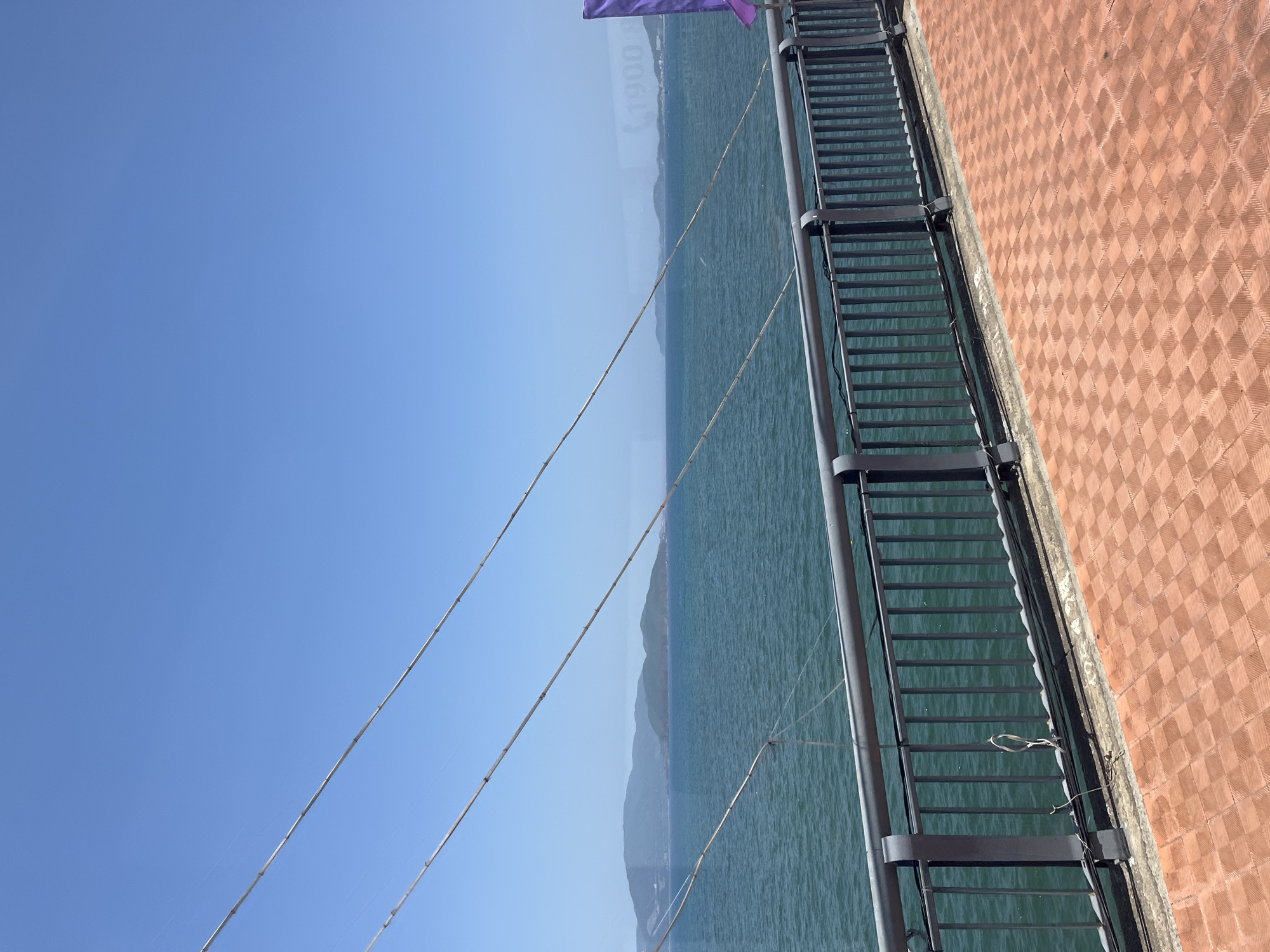 The image depicts a scenic view of Nha Trang Bay, Vietnam, from what appears to be a bridge or elevated walkway. The foreground features a textured orange tiled pathway and a dark gray metal railing, separating the walkway from the expansive turquoise ocean. Several thin, light-colored cables are stretched across the frame against a clear blue sky, suggesting the structure might be a suspension bridge or part of a larger infrastructure. In the mid-ground and background, the ocean stretches out to meet a distant, hazy coastline characterized by rolling green hills and islands. A small purple flag is visible on the right side of the frame, adding a splash of color to the scene. There are no people visible in the image. The overall impression is one of tranquility and natural beauty, typical of coastal landscapes.