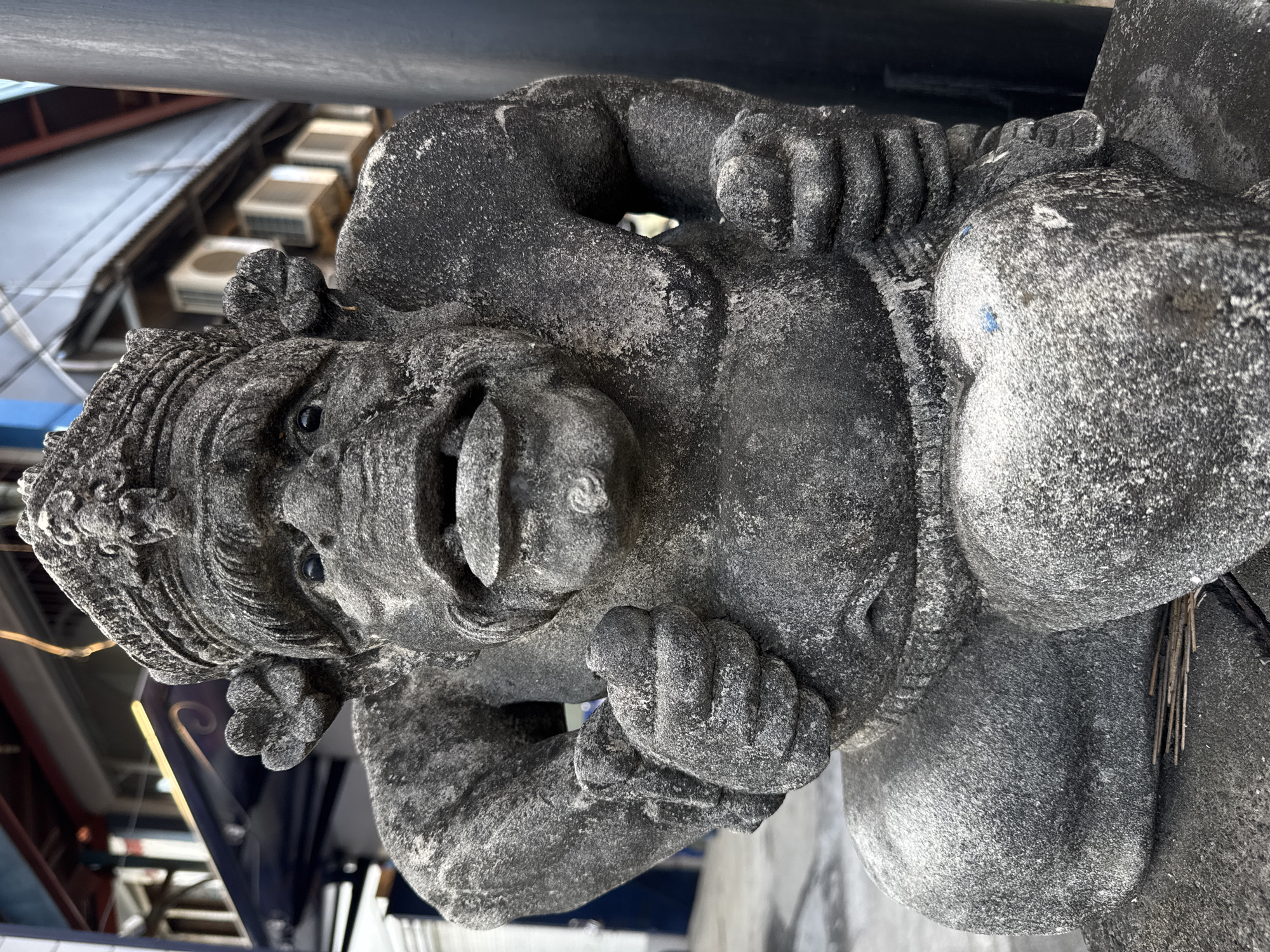 A weathered, dark gray stone statue of a grotesque, human-like figure is positioned in the foreground, filling most of the frame. The figure has a large head with a crown, wide-open mouth revealing teeth, and large ears. Its body is stout with prominent chest muscles. It is kneeling or squatting, with its hands clenched. The statue appears to be placed outdoors on a paved surface, with a dark pole visible to the right. In the blurred background, there is a building with windows and air conditioning units. The statue is located in Kuta, Indonesia.