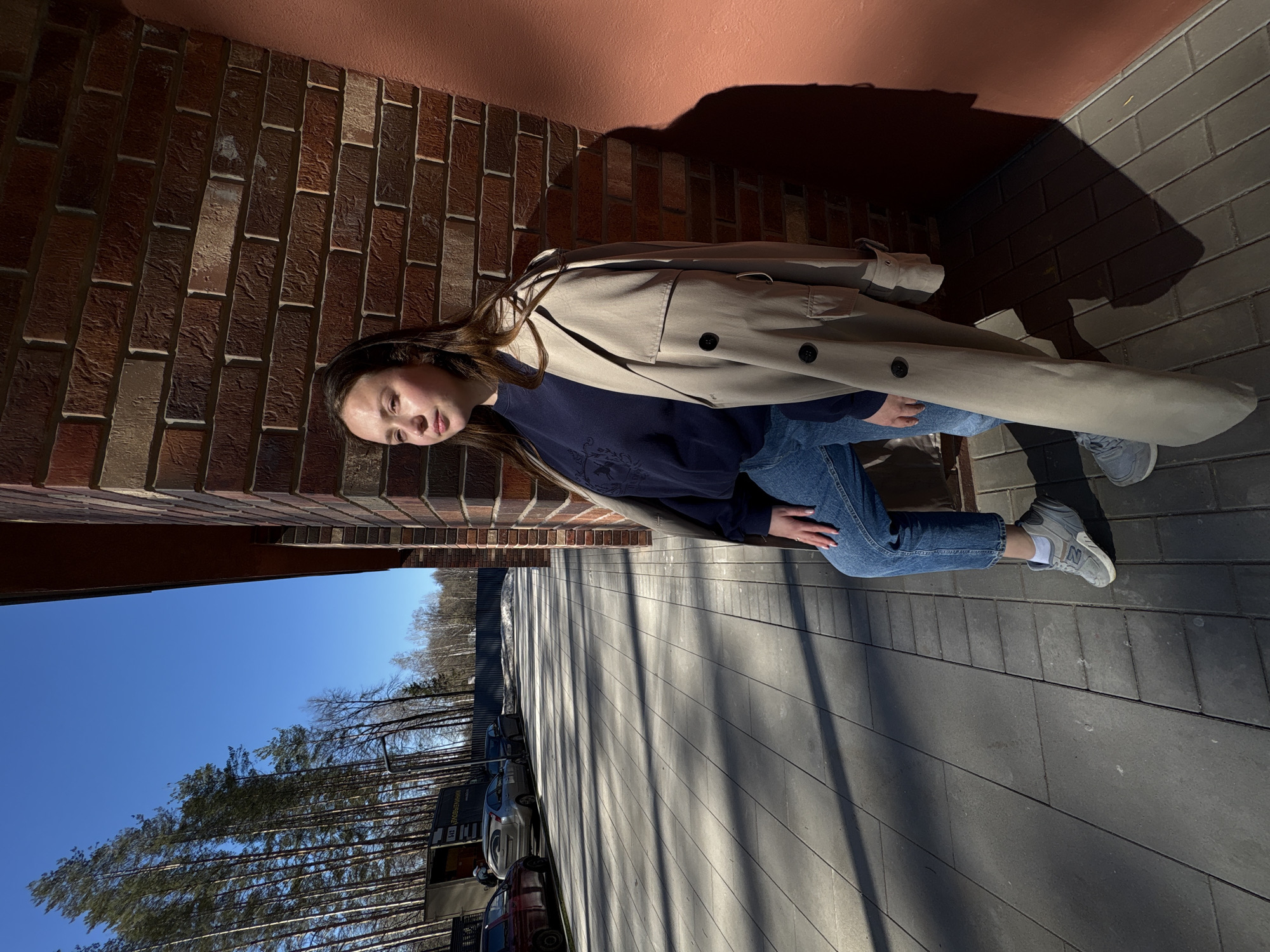 A young woman lies on a paved area next to a brick building, wearing blue jeans, a dark blue sweatshirt, a tan trench coat, and grey sneakers. She is positioned between the brick wall of the building and the edge of a paved area. A shadow is cast on the pavement next to her. A row of parked cars and trees are visible in the background beneath a clear blue sky. The scene is located in Izhevsk, Russia.