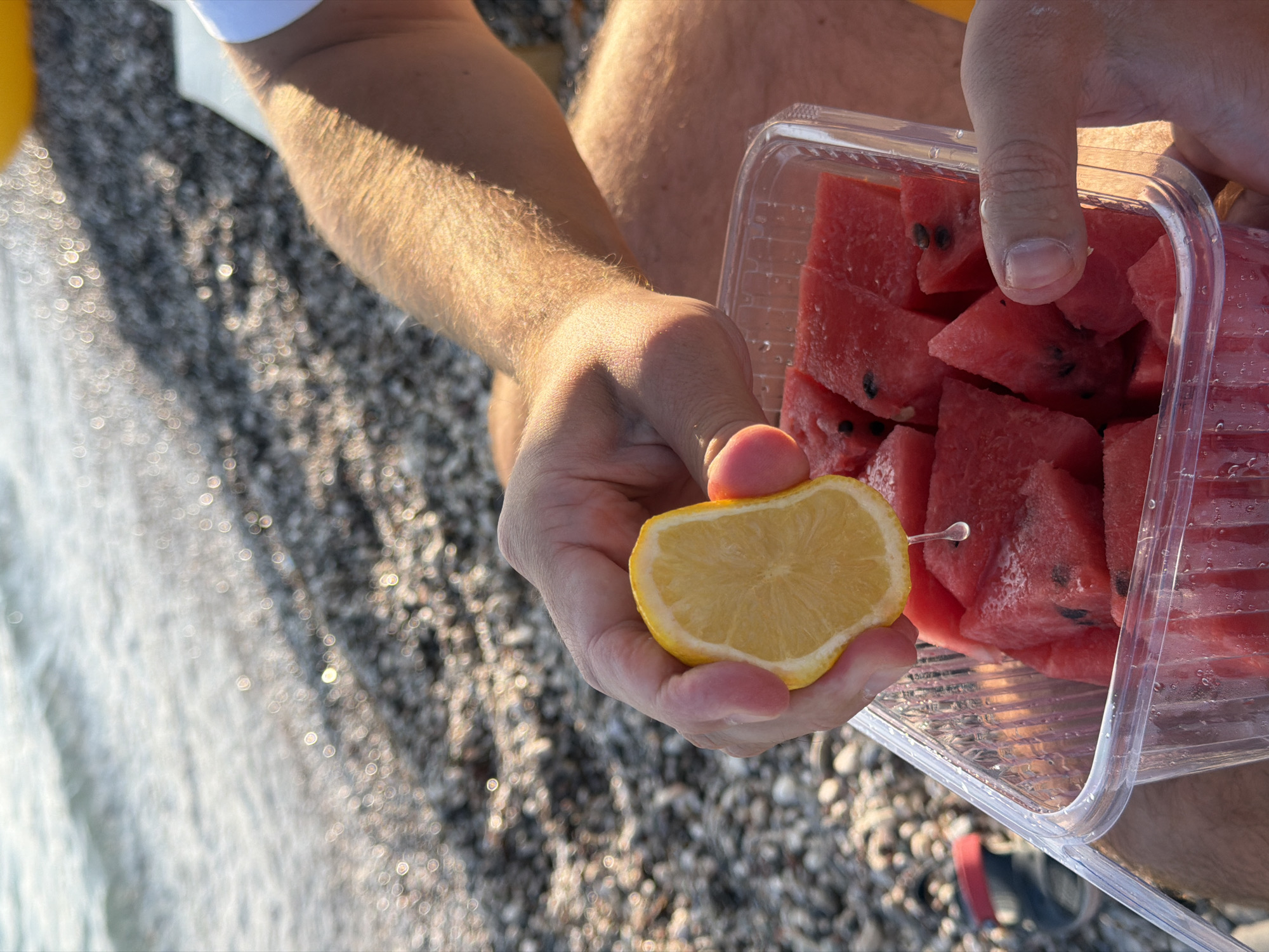 A person is holding a slice of lemon over a clear plastic container filled with cubed watermelon. The person's hand is squeezing the lemon, causing a drop of juice to fall into the watermelon. The scene takes place on a pebble beach, with the sea visible in the background. The lighting suggests it is late afternoon or early evening. The location is likely Budva, Montenegro, a popular coastal city known for its beaches.