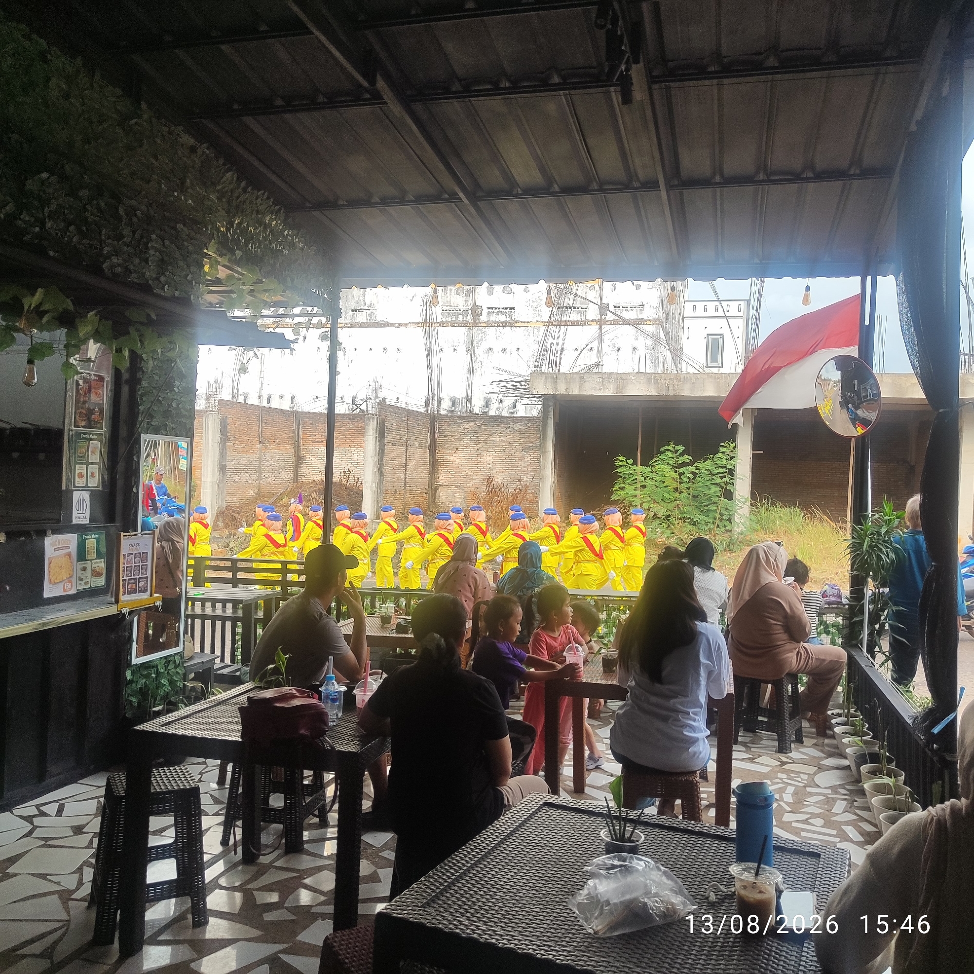 The image shows an outdoor seating area of a cafe with people dining and observing a parade of figures in yellow uniforms and blue hats. The figures appear to be mannequins or performers. In the background, there is a large white building and a brick wall. An Indonesian flag is visible in the upper right corner. The scene is lit by natural light. The cafe appears to be located in Buding, Indonesia, based on the provided information.