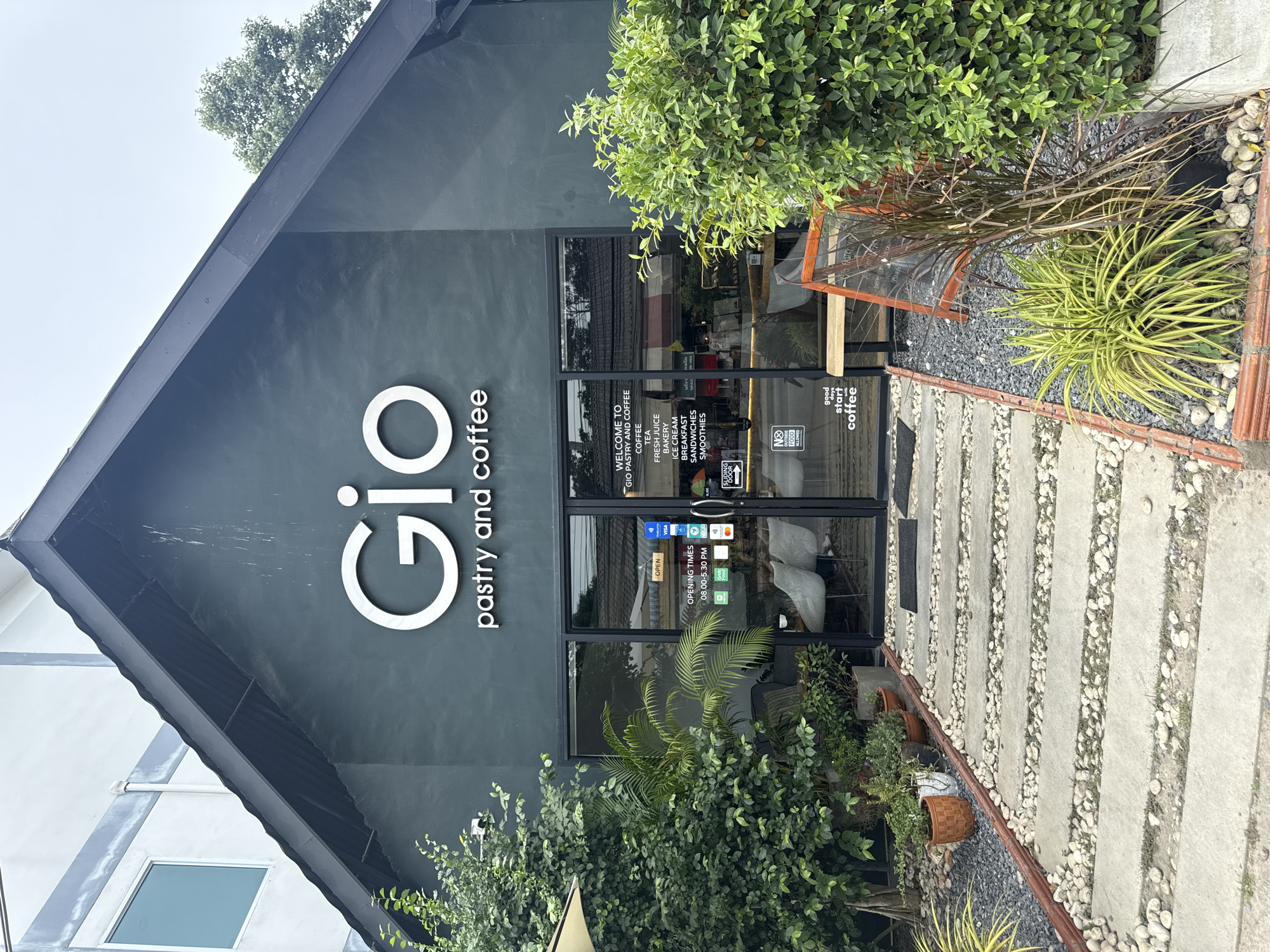 The image shows the exterior of "Gio Pastry and Coffee," a cafe located in Baan Bo Phut, Thailand. The cafe has a modern, dark green facade with a prominent white "Gio" logo above the entrance. The entrance consists of large glass doors, allowing a view into the interior where tables and chairs are visible.
Several signs are affixed to the glass doors. One sign, in English, reads "WELCOME TO GIO PASTRY AND COFFEE" and lists offerings such as "COFFEE," "TEA," "FRESH JUICE," "BAKERY," "ICE CREAM," "BREAKFAST," "SANDWICHES," and "SMOOTHIES." Another sign indicates "OPENING TIMES 08.00-5.30 PM" with icons for GrabFood and Foodpanda. Payment method logos such as Visa, Mastercard, and UnionPay are also displayed. A smaller "NO OUTSIDE FOOD ALLOWED" sign is present. A sliding door is also indicated.
Outside the entrance, a pathway made of large concrete slabs interspersed with white pebbles leads to the doors. This path is flanked by gravel and various plants, including lush green bushes, potted plants, and ornamental grasses. To the right of the entrance, an A-frame chalkboard sign with a wooden frame is placed, displaying handwritten text, presumably menu items or daily specials, though the text is largely illegible at this resolution. A wooden bench is situated near the sign.
The surrounding environment appears to be an urban or semi-urban area, with the suggestion of other buildings and trees in the background. The sky is overcast. The overall impression is of a welcoming and well-maintained cafe.