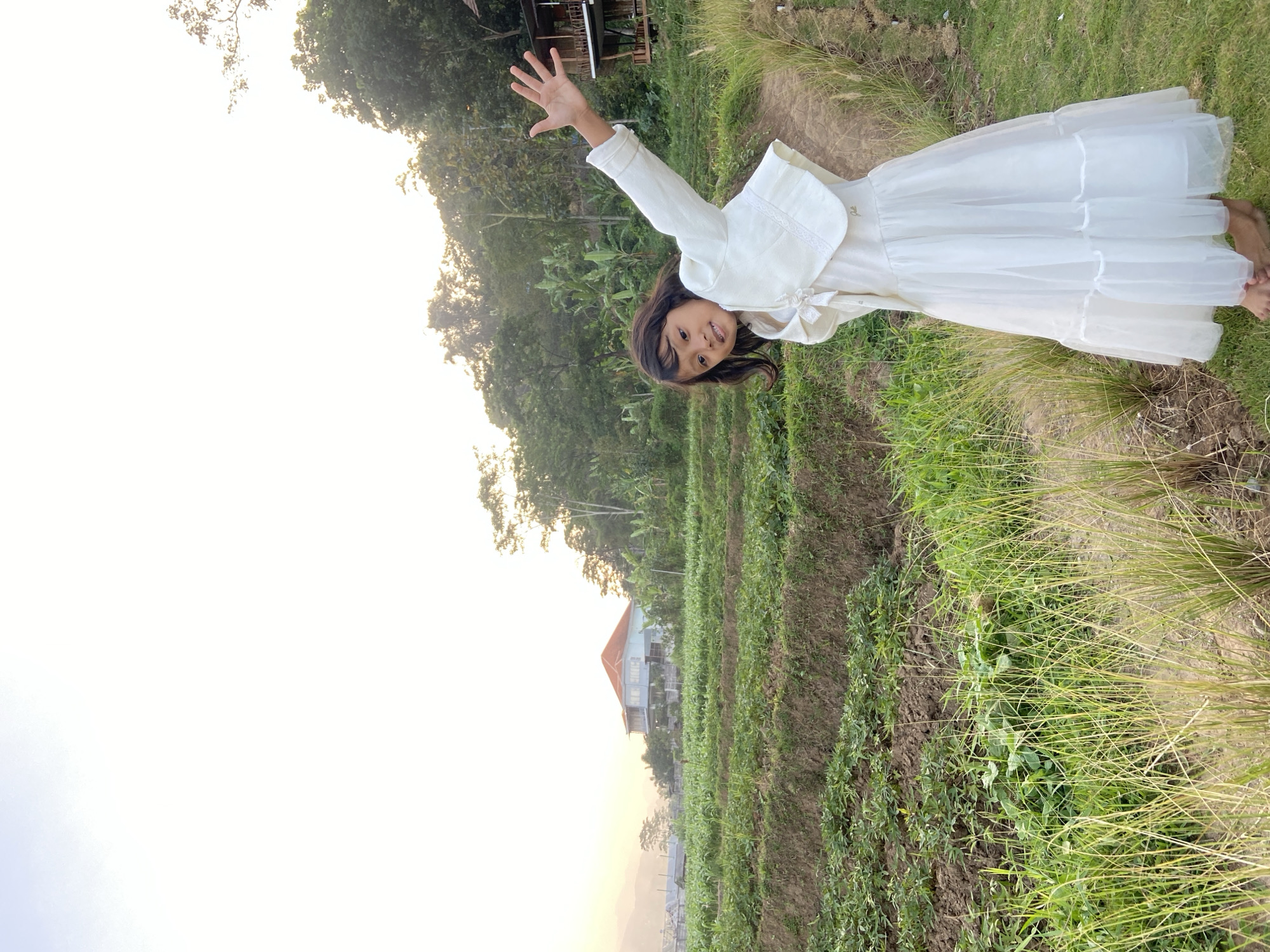 A young girl wearing a white dress and jacket stands in front of a rural landscape. The girl is positioned on the right side of the frame, with her left arm raised and hand open, as if waving. Her dress is long and appears to be made of a sheer material, with layers at the hem. The jacket is short and white, with lace trim.
The background features tiered fields of green crops, characteristic of agricultural terraces. A house with a red roof is visible in the middle distance, situated on a hill among trees. The sky above is bright and overcast, suggesting either dawn or dusk. The overall setting is natural and appears to be in a village or rural area, possibly in the city of Kesiman, Indonesia, given the vegetation and architectural style.